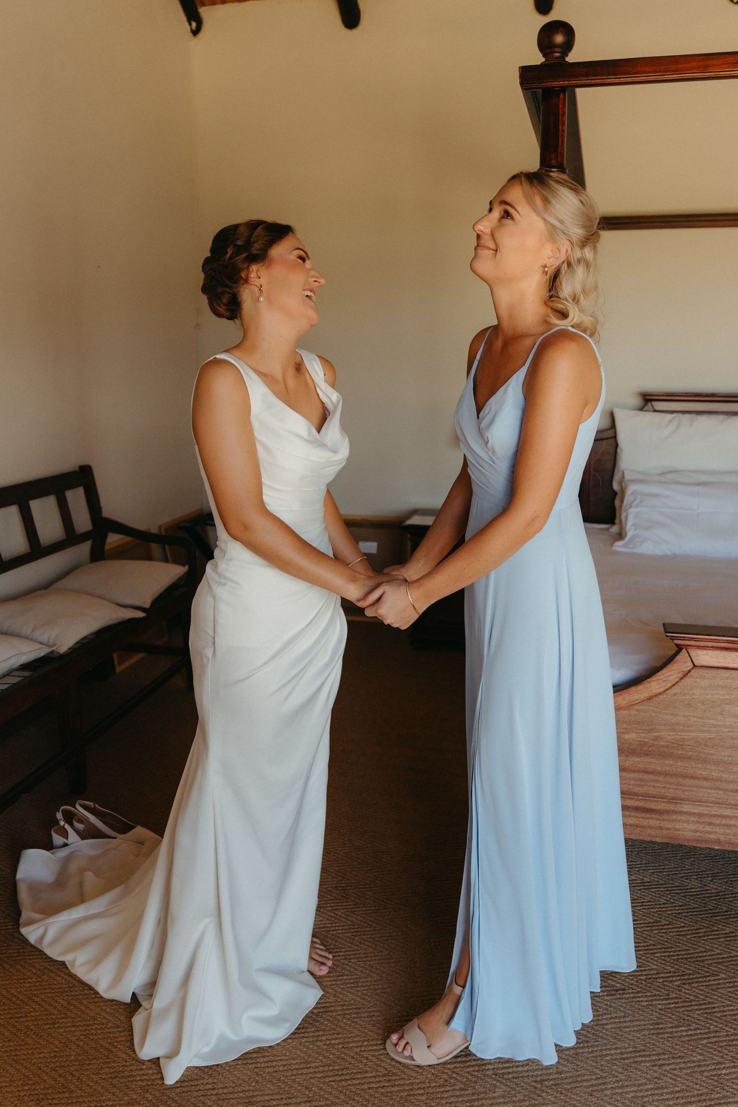 Two people in white and light blue dresses share an emotional moment while getting ready in a sunlit room.