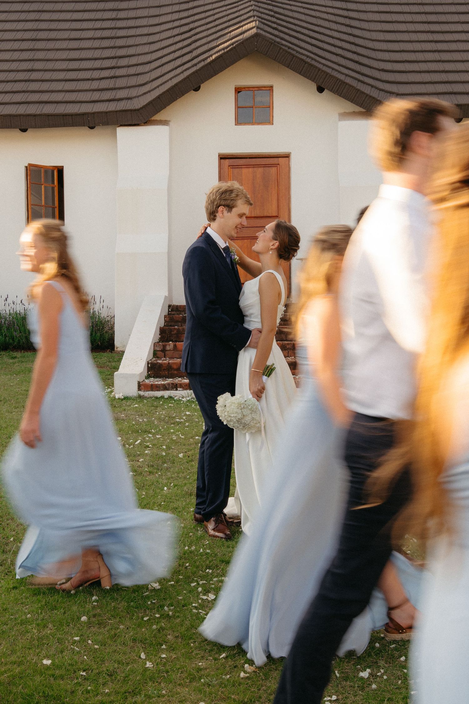 Motion blur captures wedding party walking together outdoors near white building walls in formal attire.