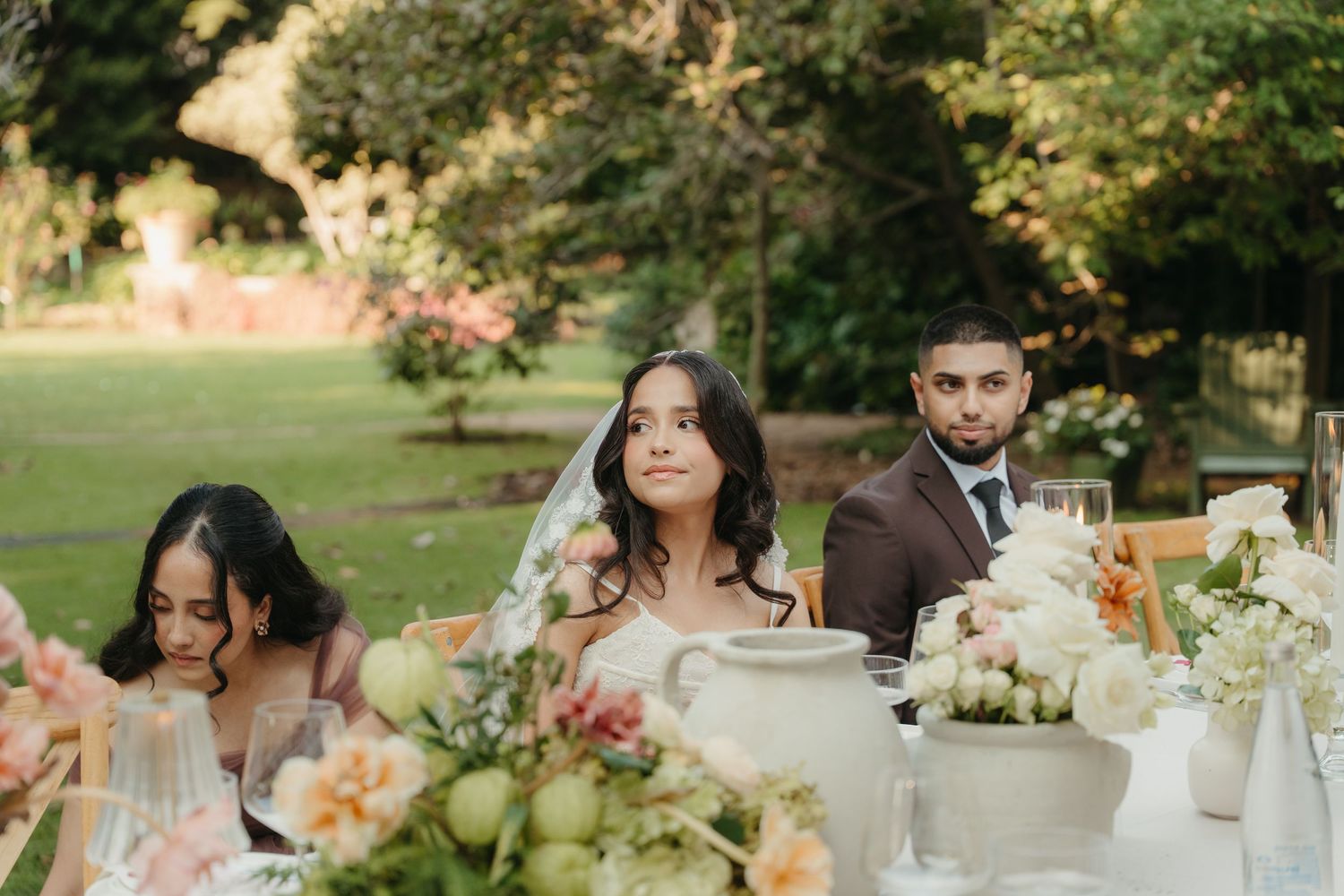 Bride and groom seated at elegant outdoor wedding reception table with floral arrangements and lush green garden backdrop.