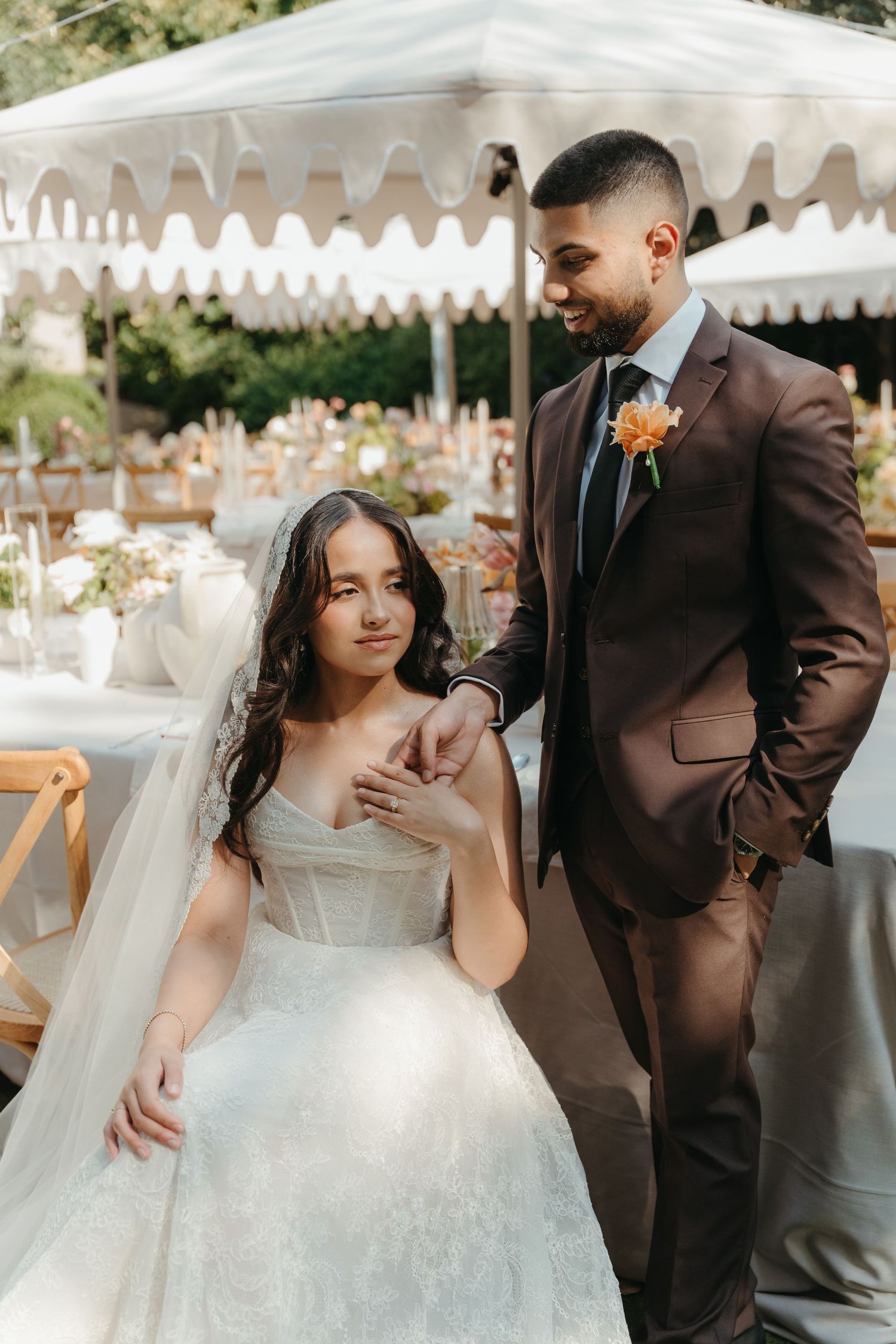 Bride in white gown sits as groom in brown suit stands beside her at an outdoor wedding reception under a white tent.