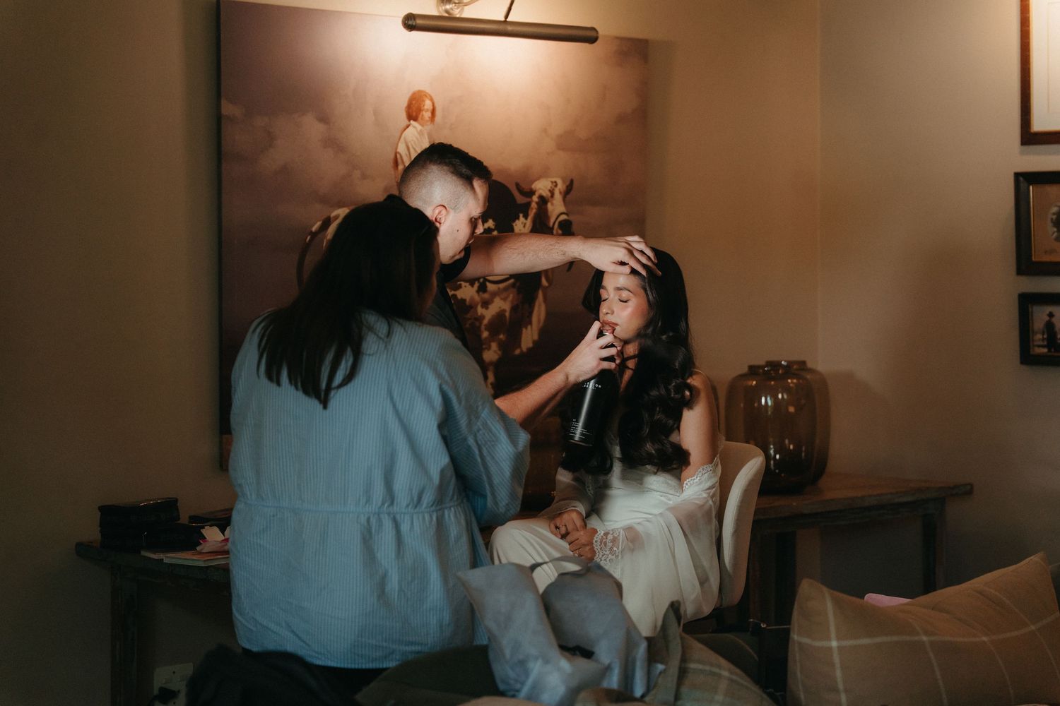 Bride getting hair and makeup done by two stylists in a warmly lit dressing room at Beechwood Gardens before her wedding.