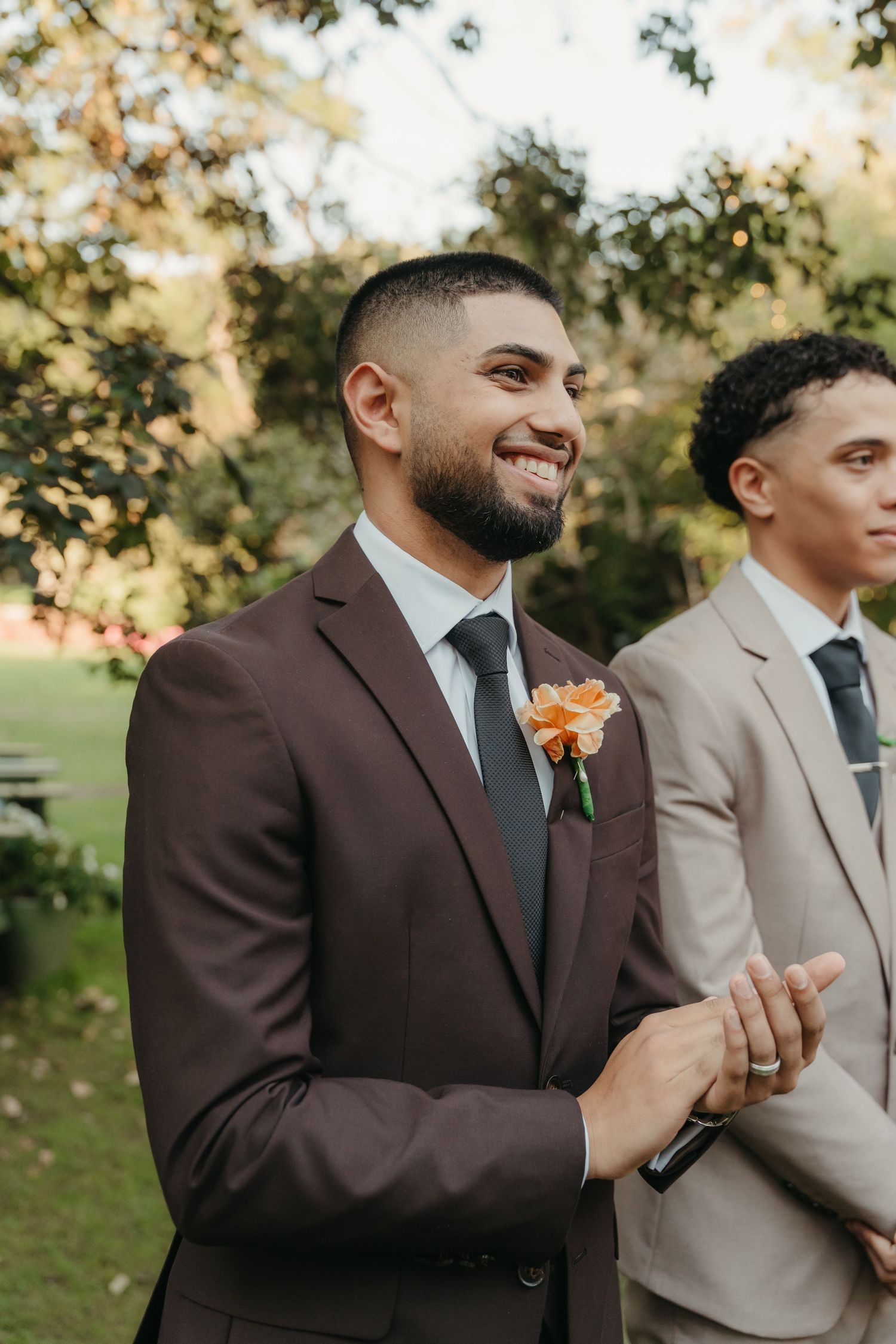 Smiling groom in dark brown suit with orange boutonniere stands outdoors at wedding ceremony beside groomsman in tan suit.