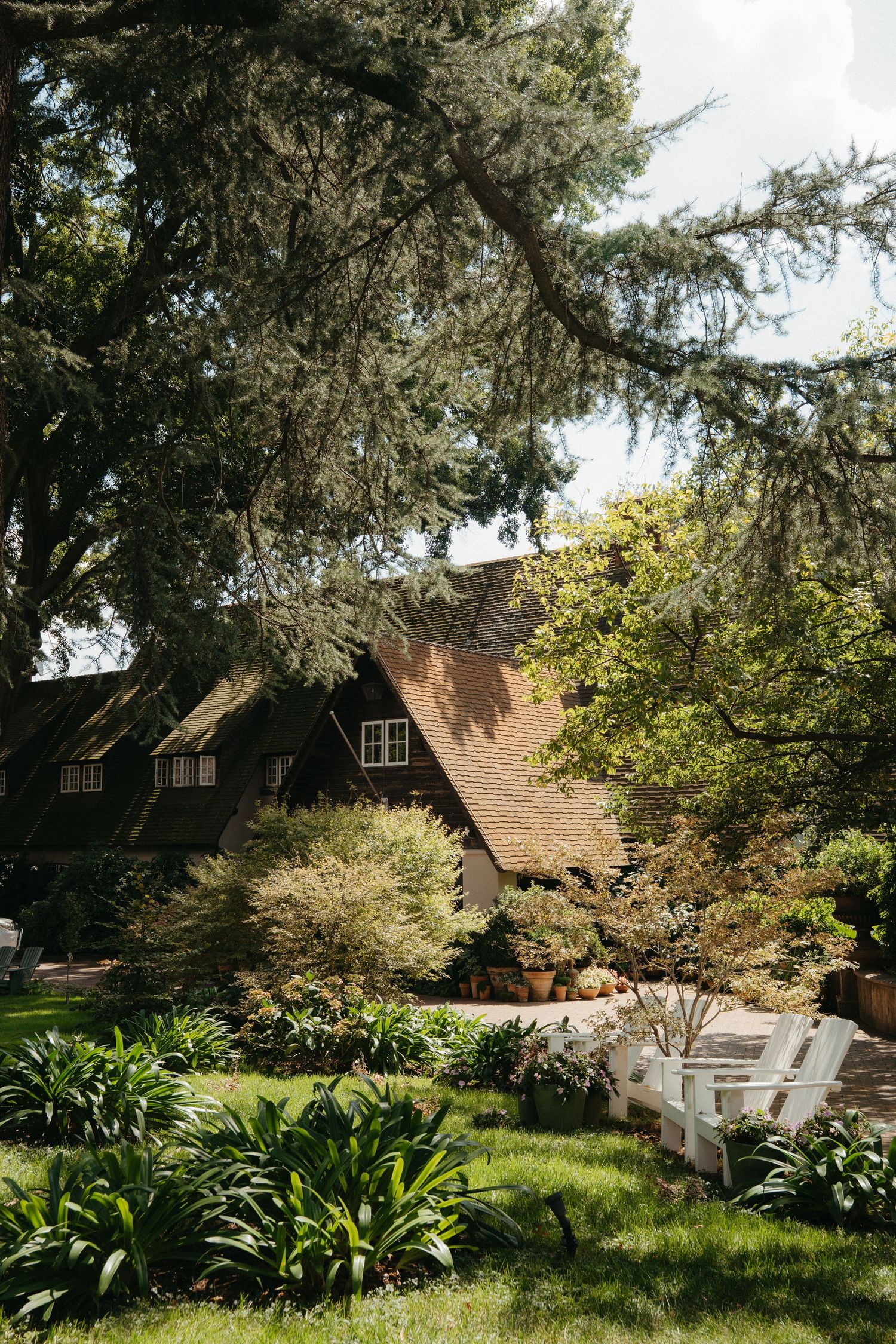 Charming cottage-style Beechwood Gardens with brown roof surrounded by lush green gardens, mature trees, and white Adirondack chairs.