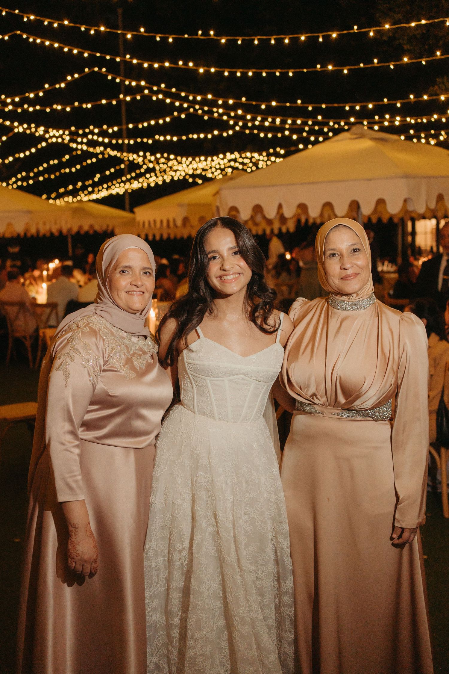 Bride in white corset gown poses with two women in champagne hijabs under string lights at outdoor wedding reception.