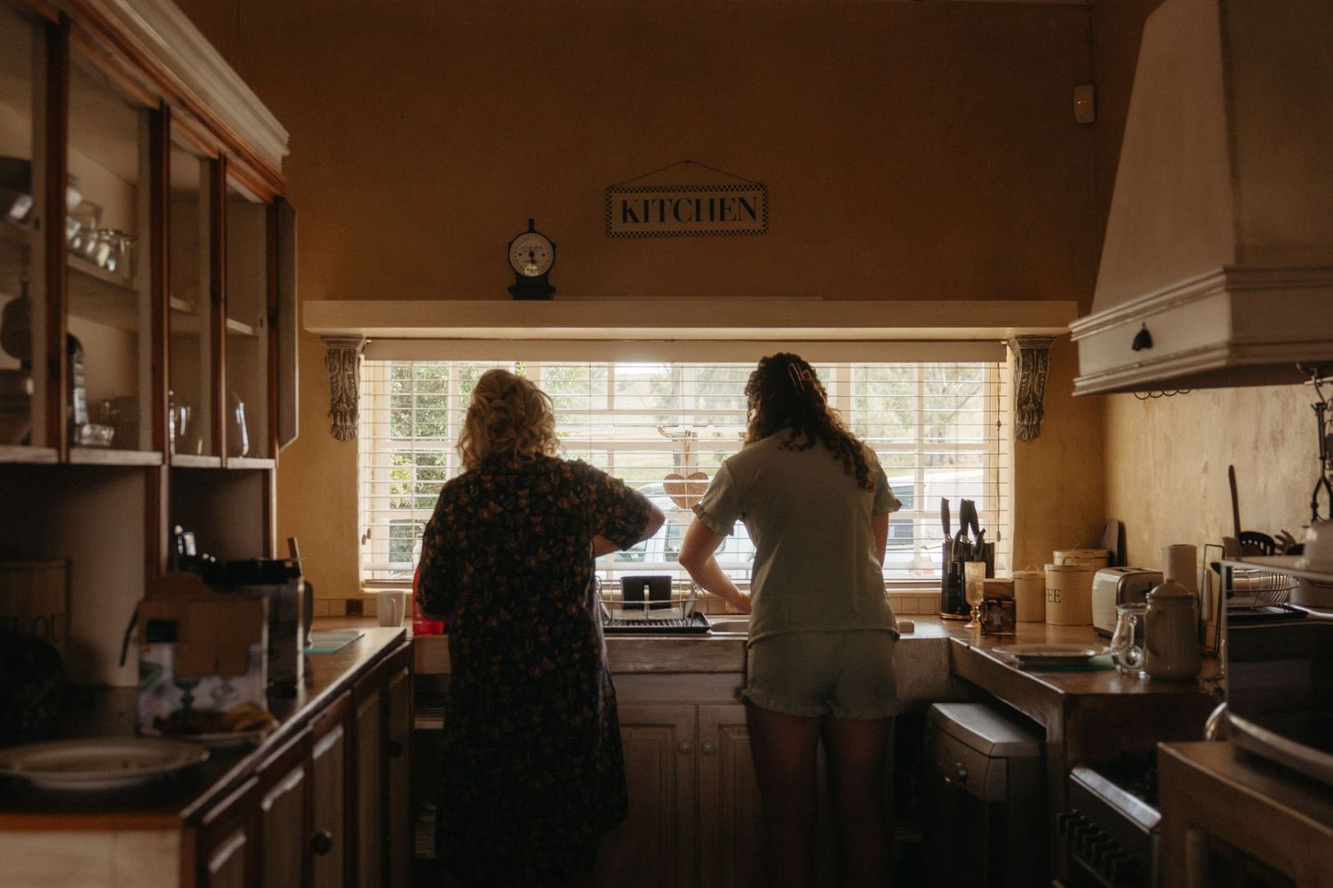Two women stand together in a warm vintage kitchen looking out the window.