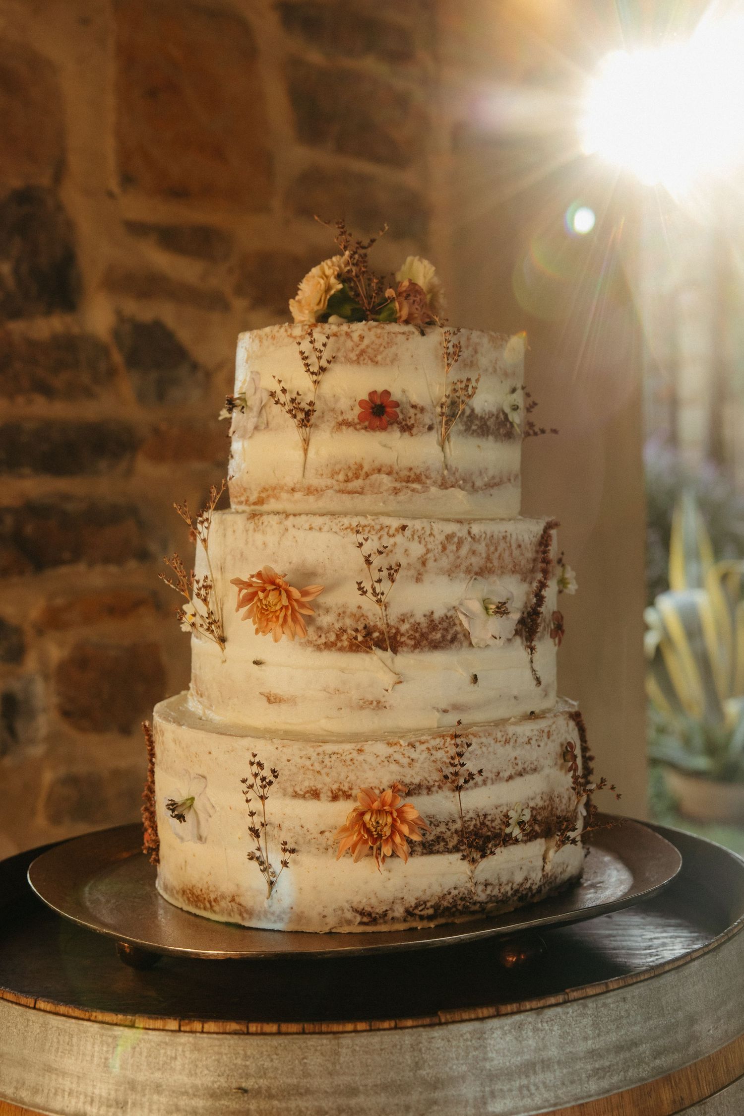 Three-tier semi-naked wedding cake decorated with dried flowers and greenery, displayed on a barrel in warm light.