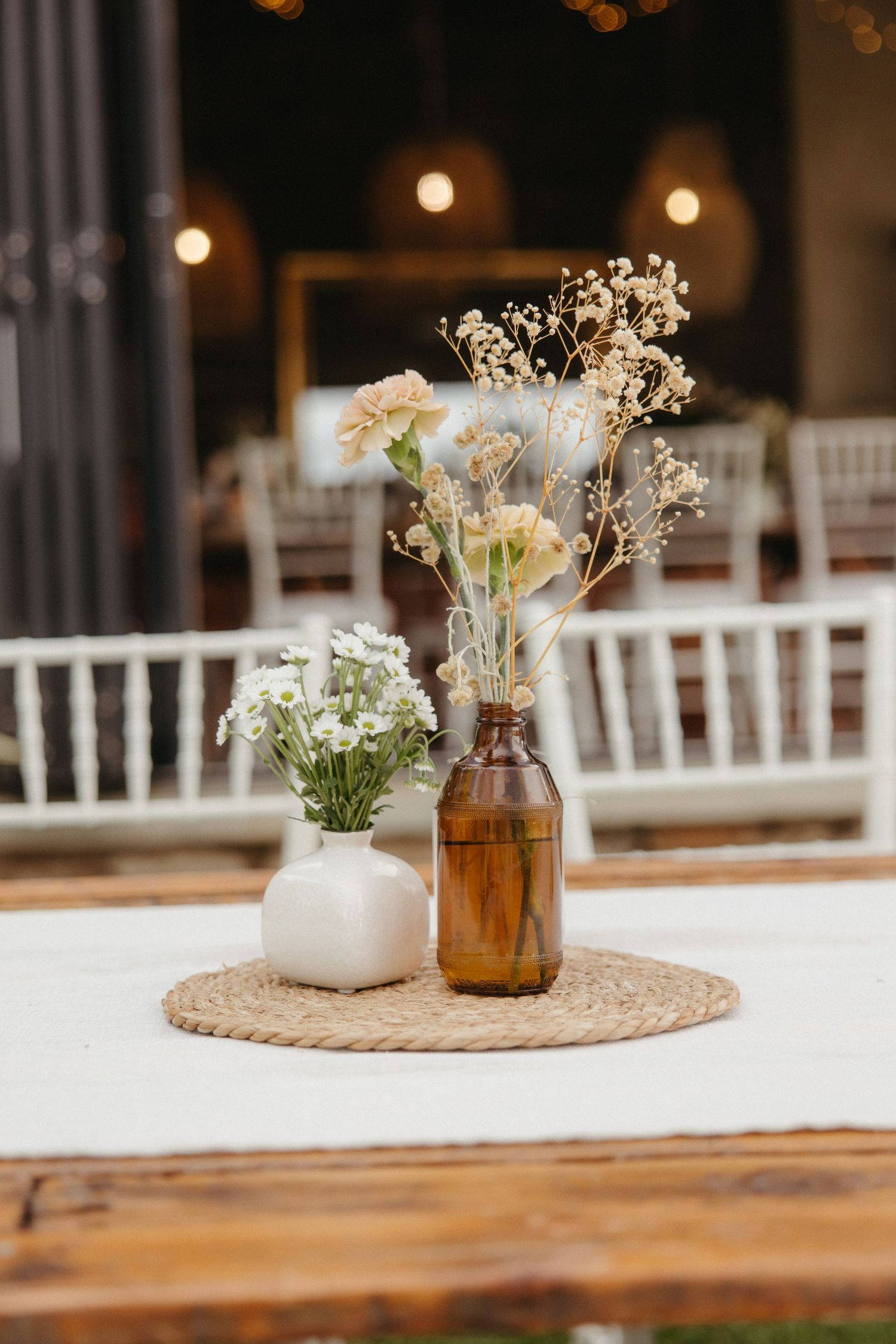 Rustic wedding table centerpiece with white ceramic vase and amber bottle holding wildflowers on woven mat.