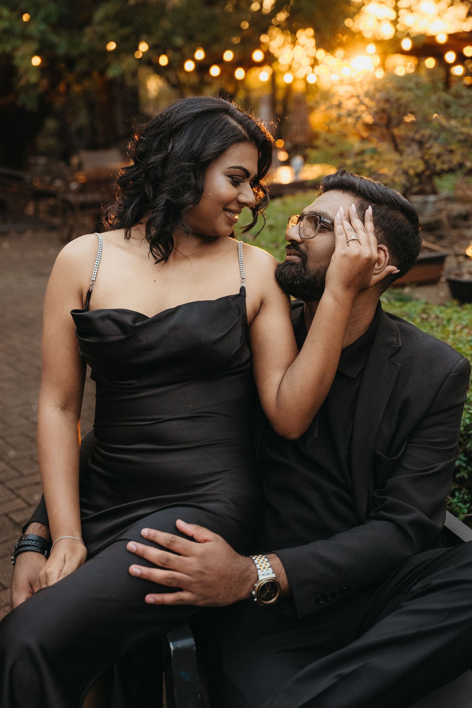 Couple in matching black formal attire sharing a romantic moment outdoors under warm string lights at sunset.