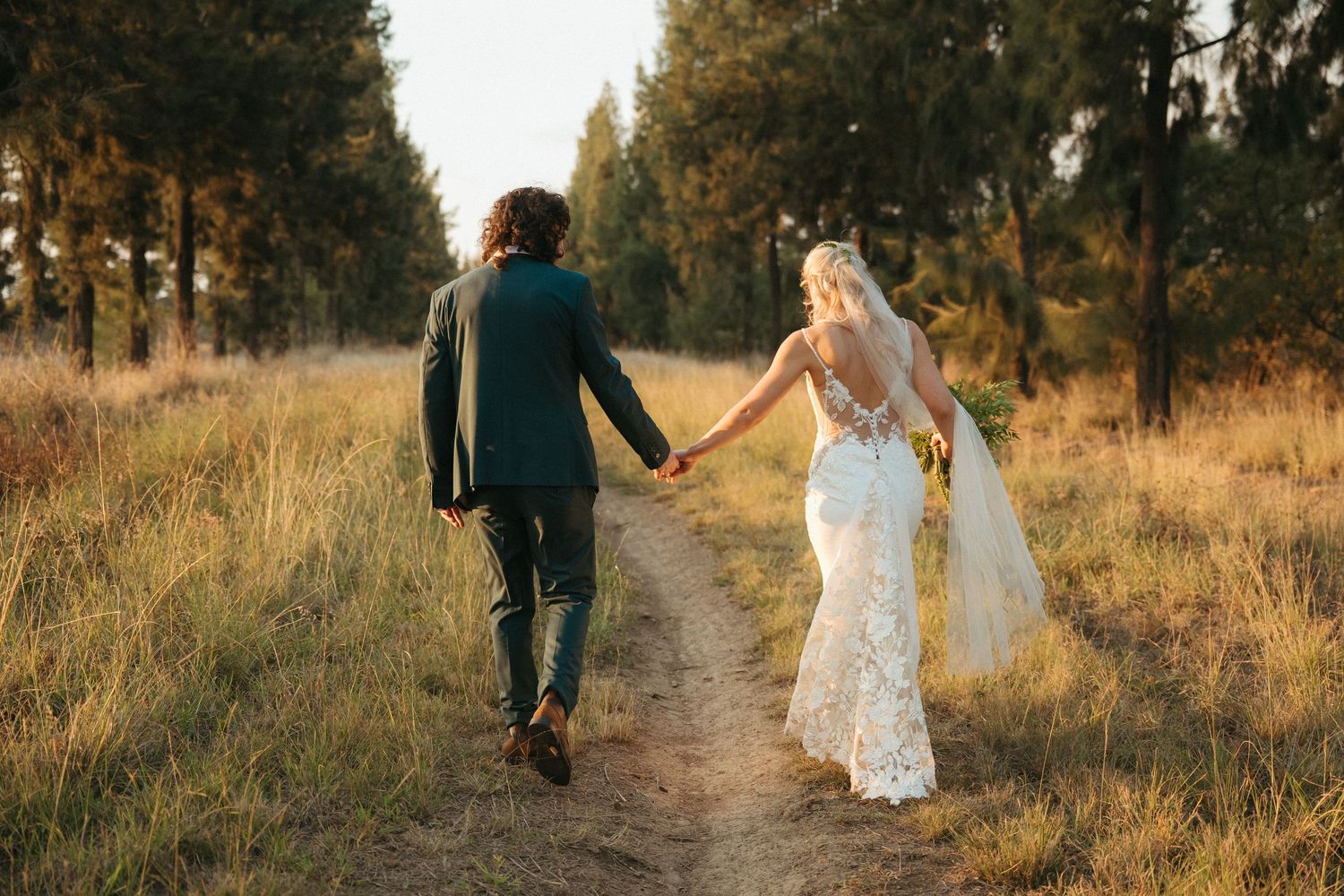 A bride and groom walk hand-in-hand down a sunlit dirt path surrounded by golden grass and tall trees.