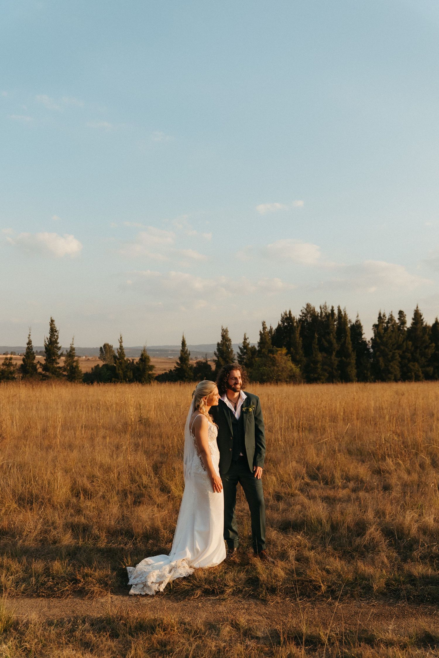 Bride and groom standing in golden field at sunset with pine trees in background on their wedding day.