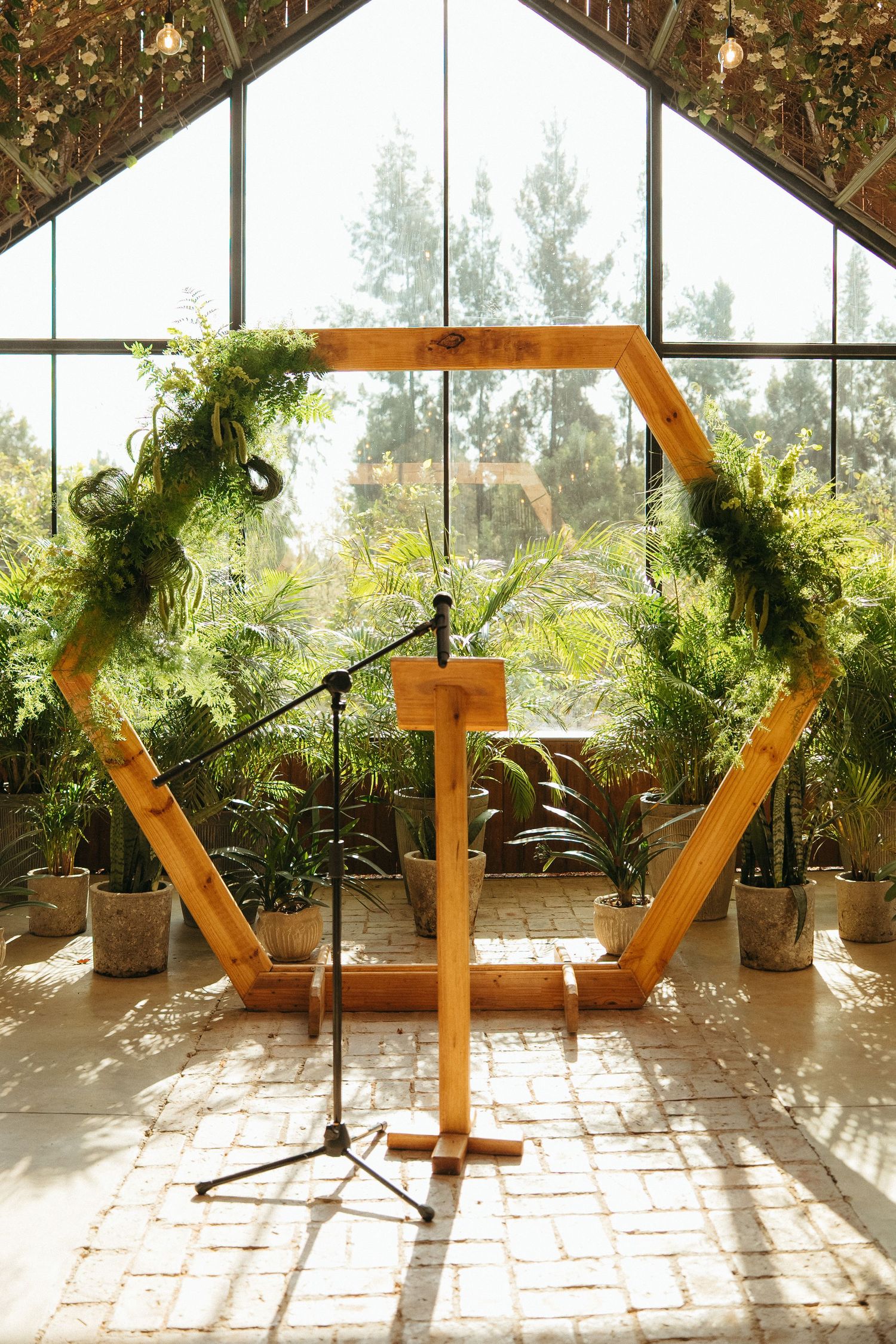 Hexagonal wooden wedding arch with greenery, podium, and microphone in a sunlit greenhouse venue.