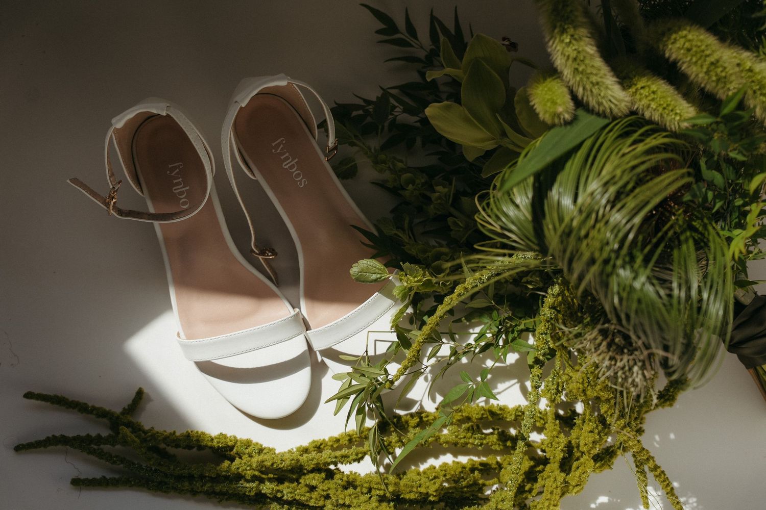 White bridal heeled sandals resting beside a lush green wedding bouquet in soft natural light.