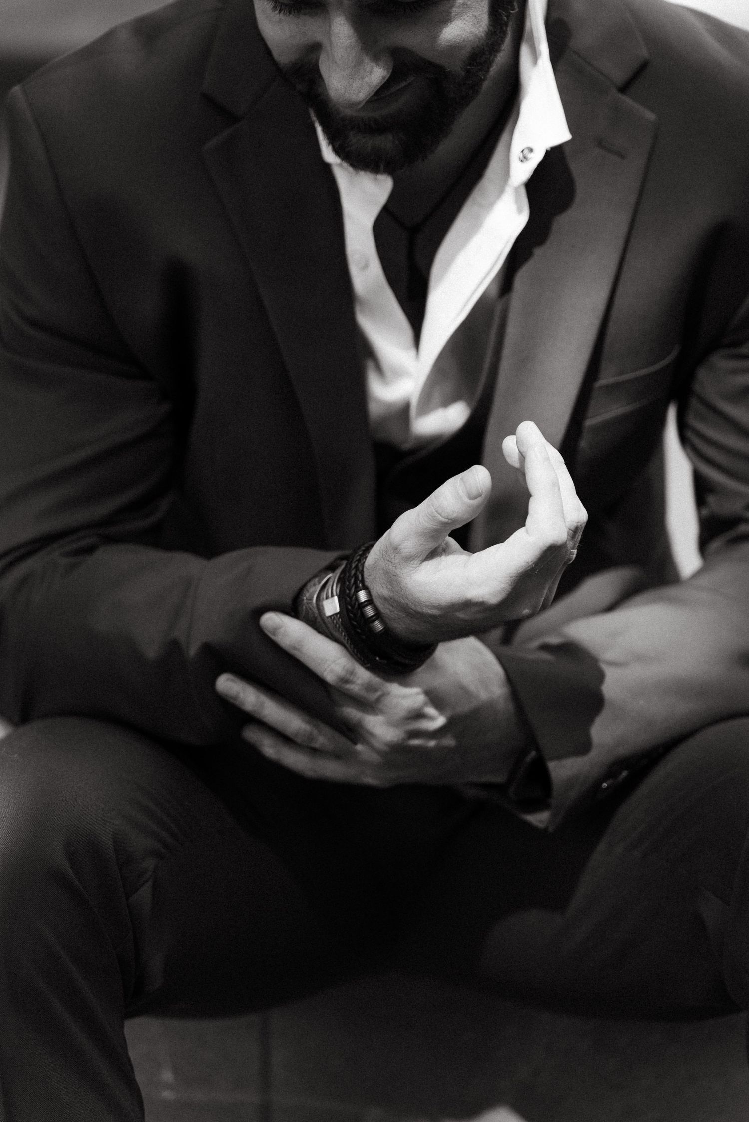 Black and white photo of a bearded man in a suit adjusting his bracelet while seated.