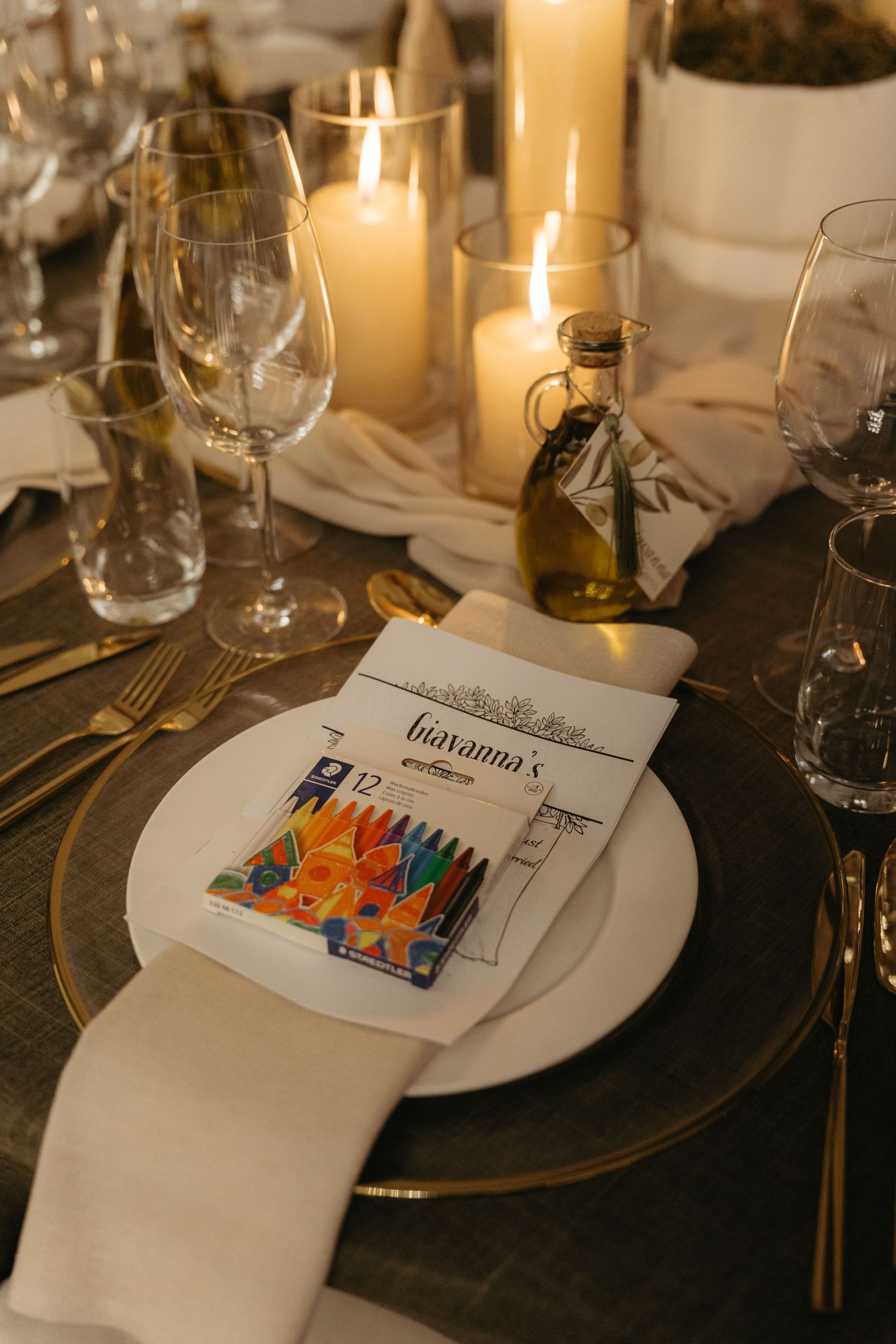Elegant wedding table setting with crayons, menu card, candles, gold cutlery, and olive oil bottle centerpiece.