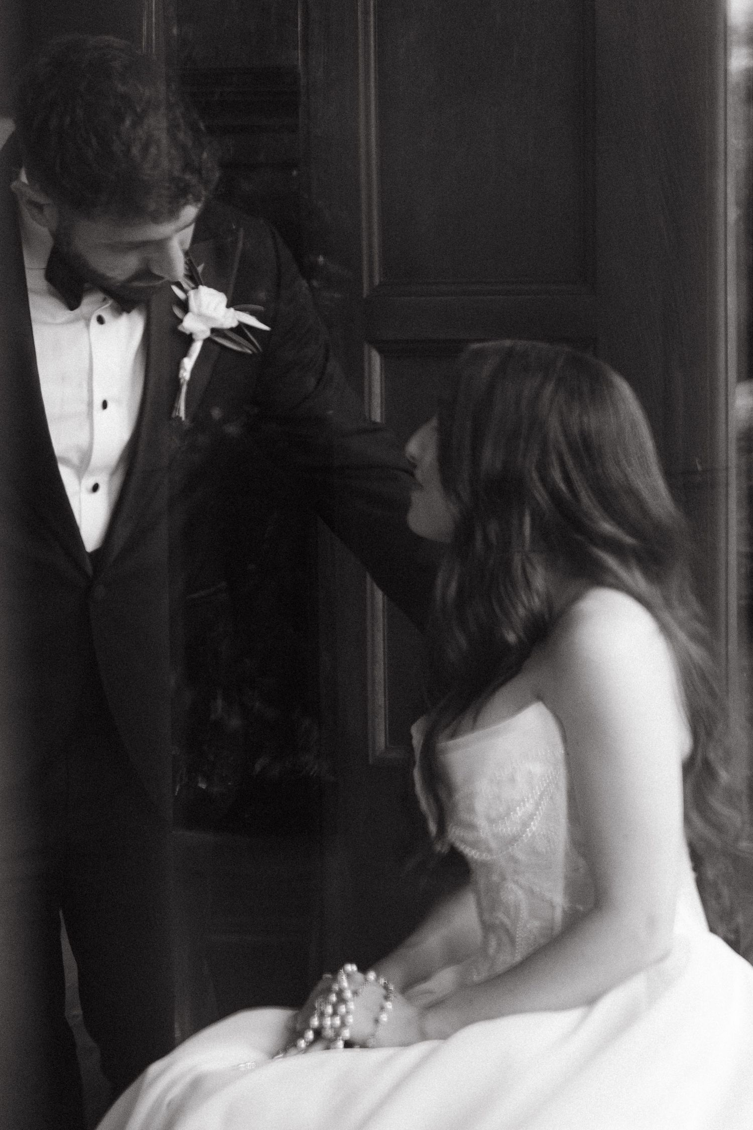 Black and white wedding photo of bride in strapless gown pinning boutonniere on groom in tuxedo.