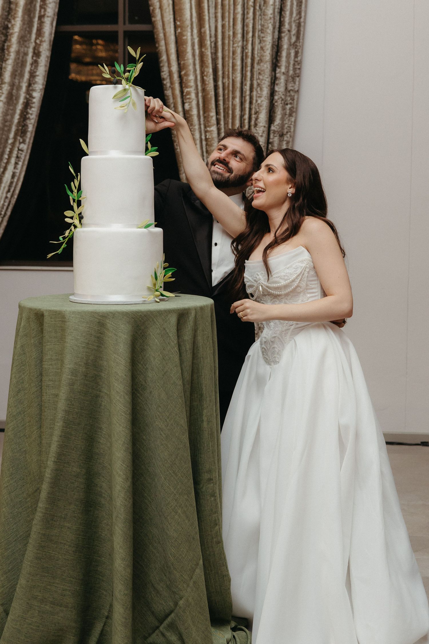Bride and groom smiling while decorating a four-tier white wedding cake with greenery on a draped table.