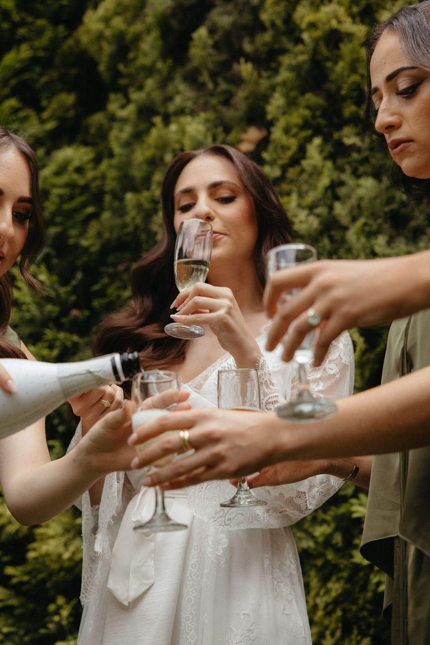 Bride in white lace robe toasting champagne with bridesmaids outdoors surrounded by lush green trees.