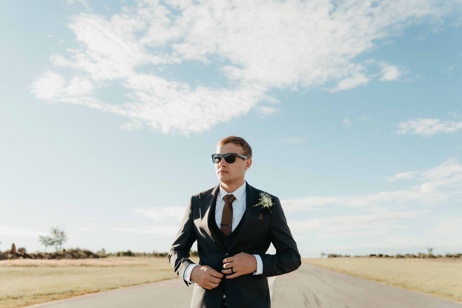A stylish groom in a dark suit and sunglasses stands on an open road under a bright blue sky.