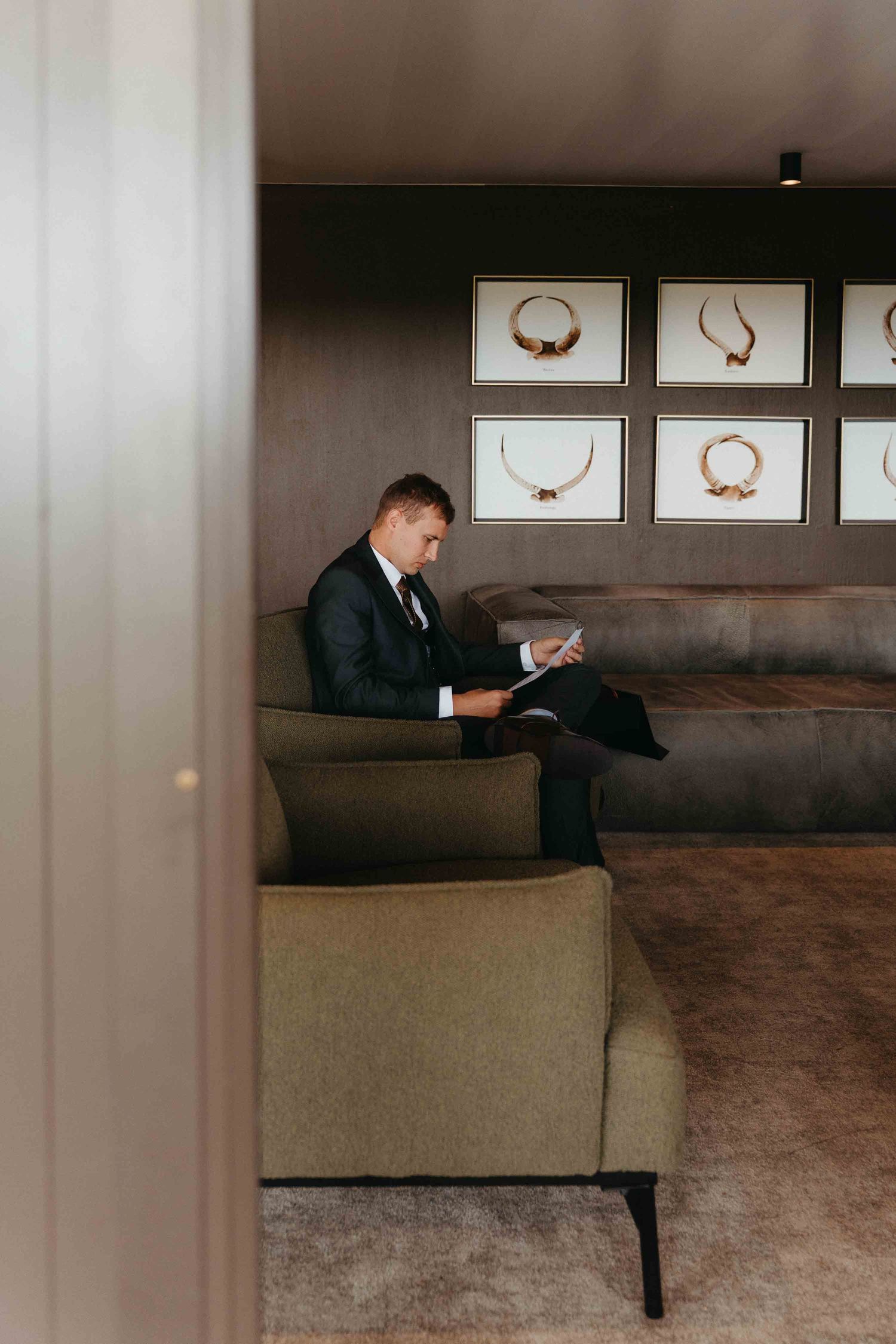 A man in a dark suit sits in a green armchair, reviewing documents in an elegant room with framed jewelry displays.