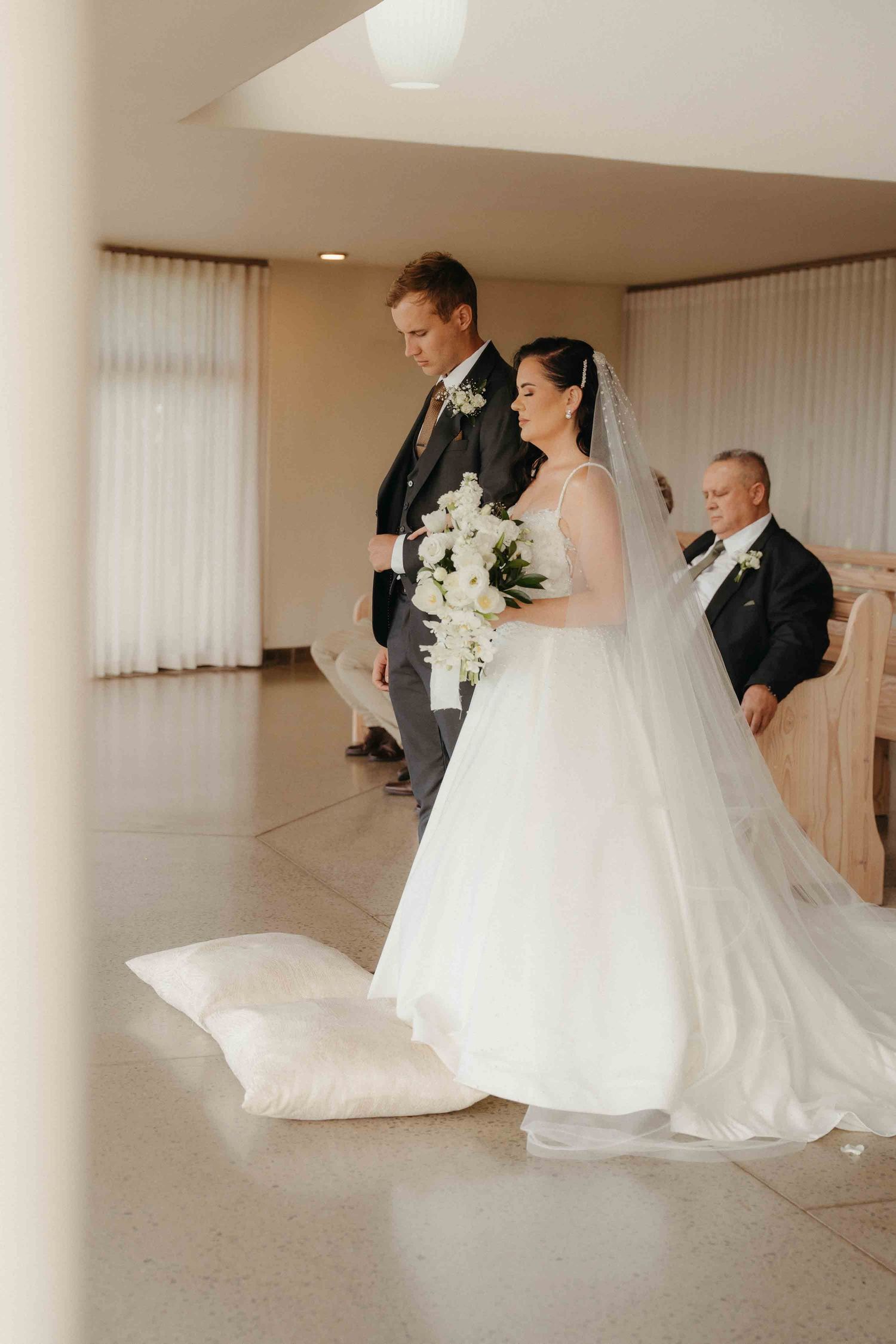 Bride in white gown holding bouquet walks with groom in gray suit during elegant indoor wedding ceremony.