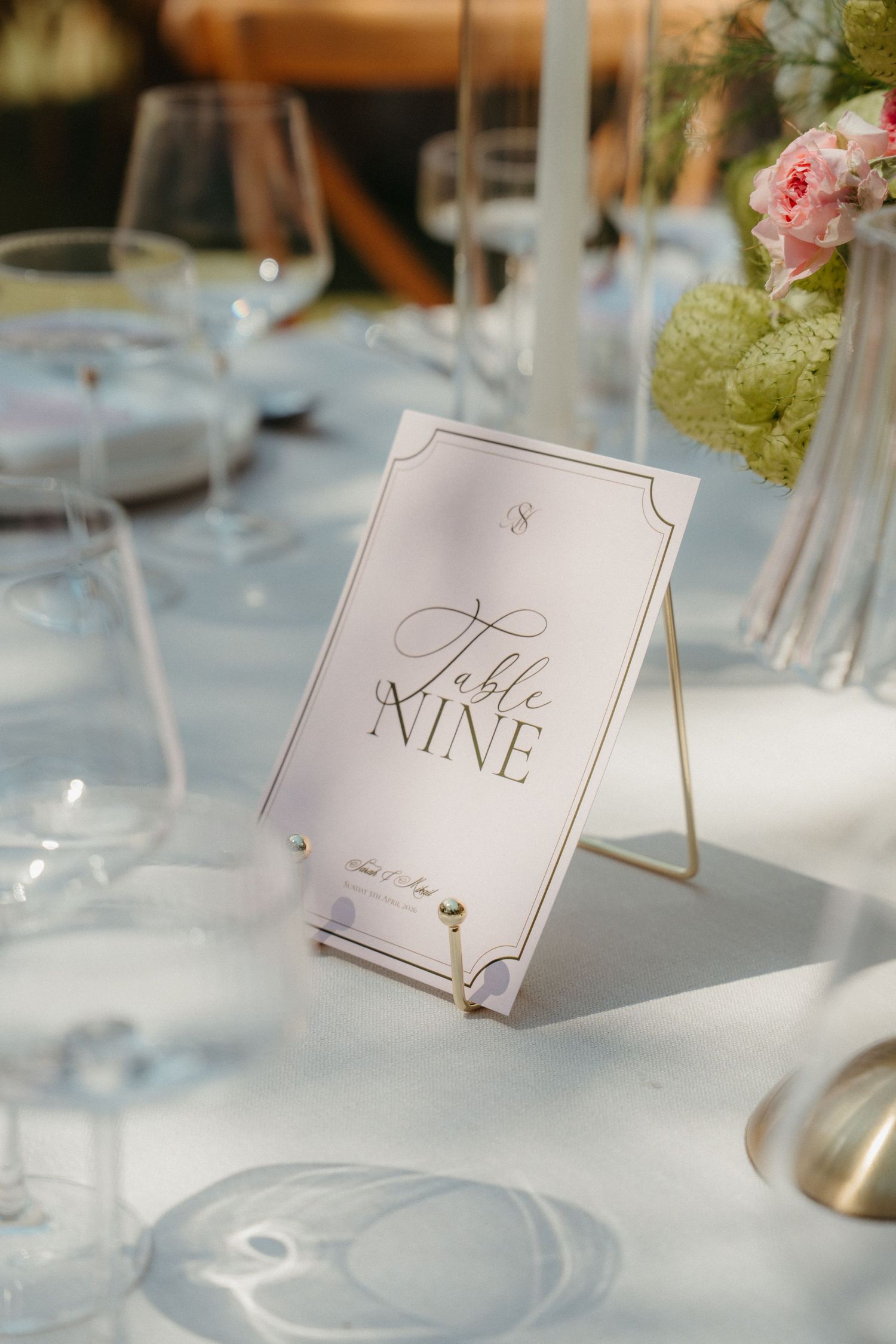 Elegant wedding table nine card in gold stand with floral centerpiece and crystal glassware on white linen.