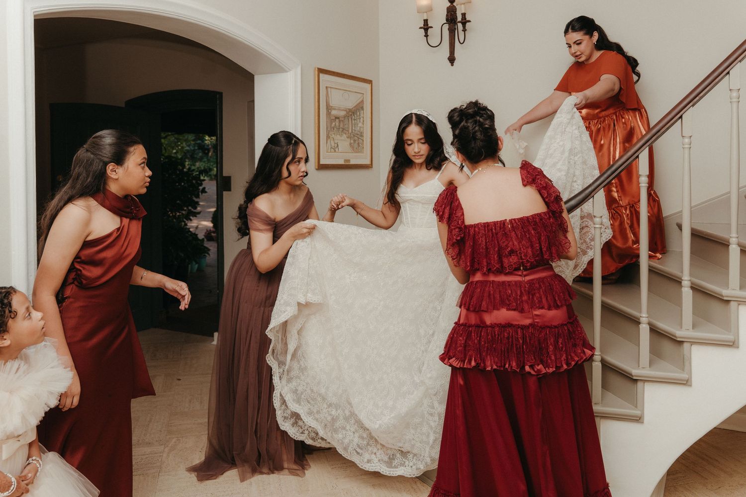 Bridesmaids in red and brown dresses help bride with white lace gown near elegant staircase indoors.