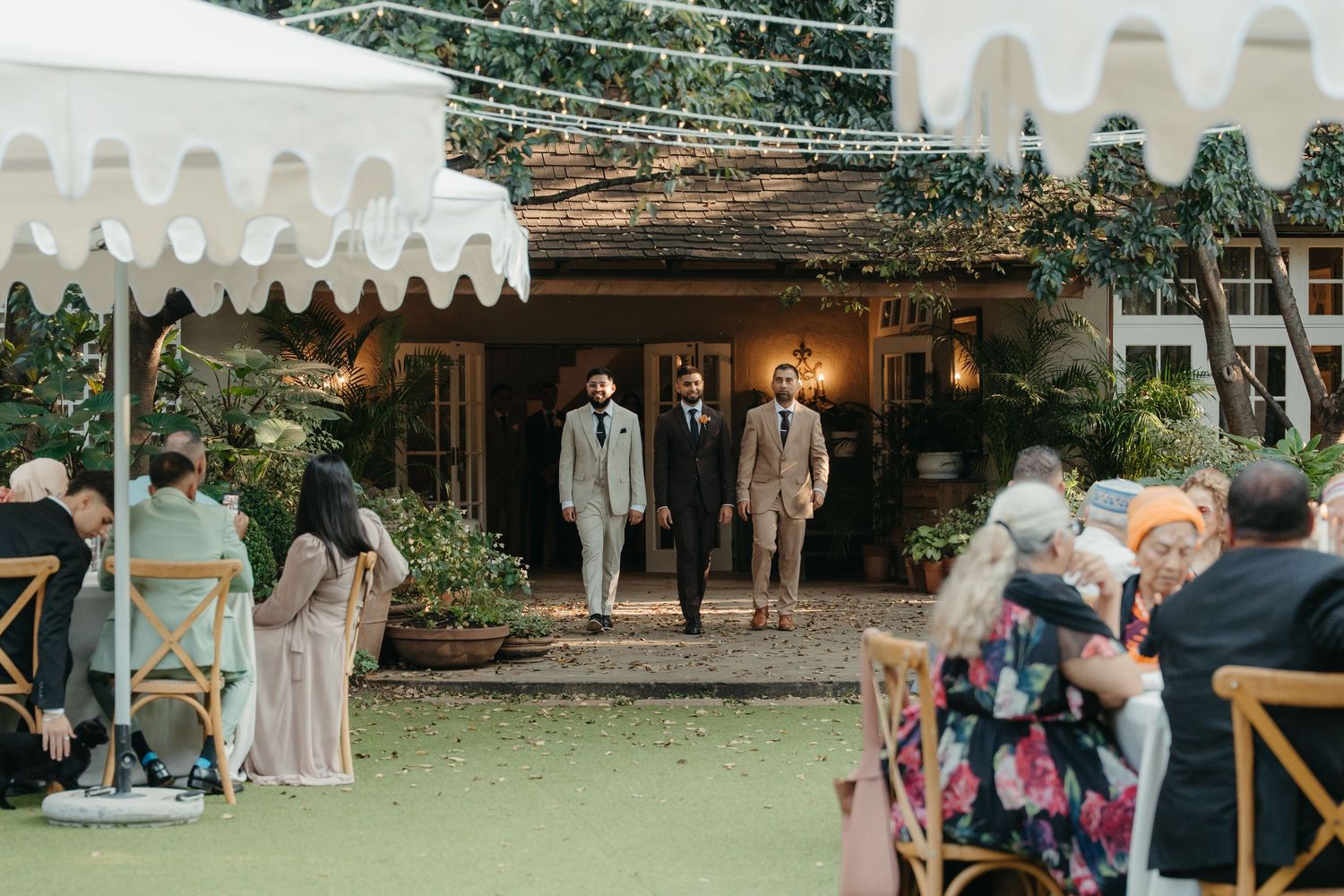 Three groomsmen in tan suits walk through a garden wedding venue entrance as guests are seated outdoors.