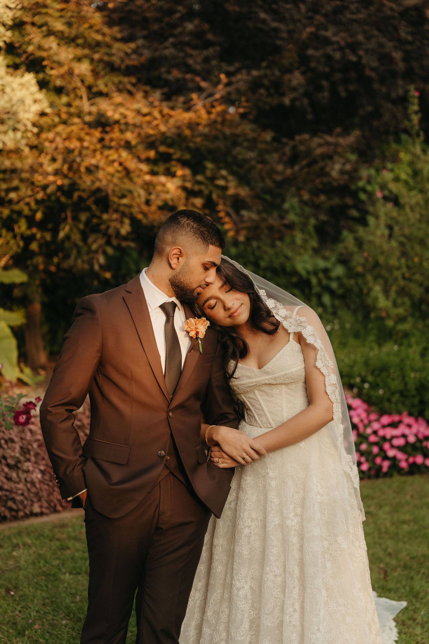 A bride in lace gown and veil leans on groom in brown suit, posing in a lush garden at golden hour.