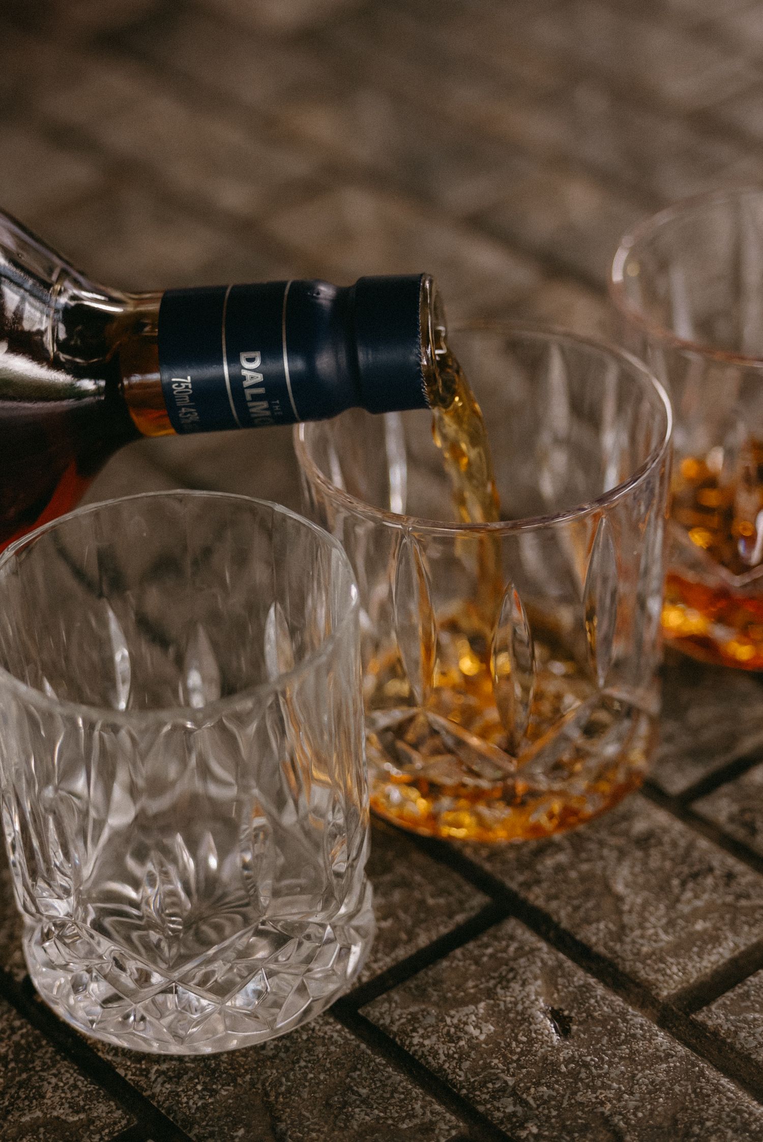 Crystal whiskey glasses being filled with amber liquid on a dark textured surface.