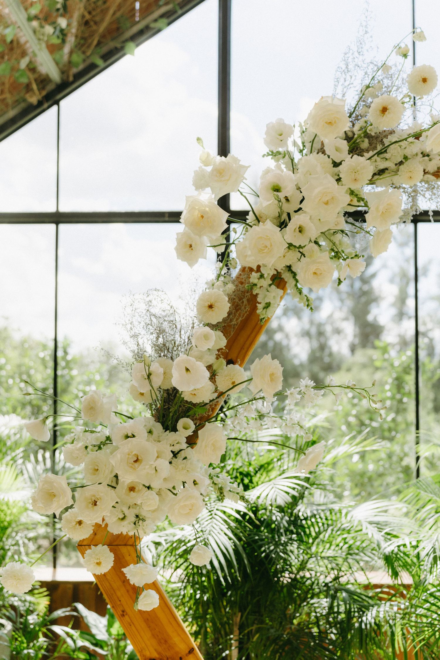 White floral arch arrangement with roses and greenery in greenhouse setting.