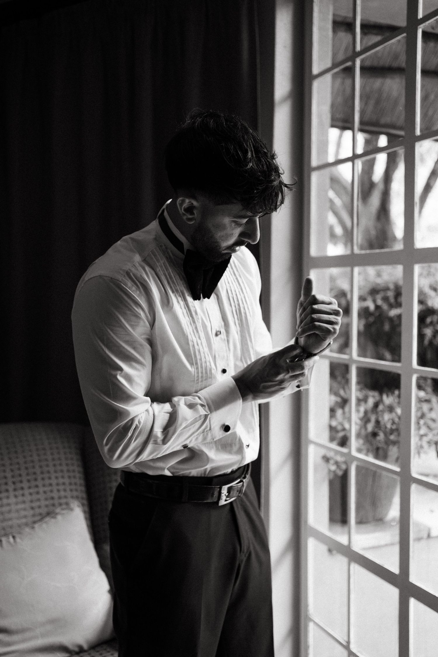 A person in a white dress shirt adjusts their cuffs while standing by a window in moody lighting.