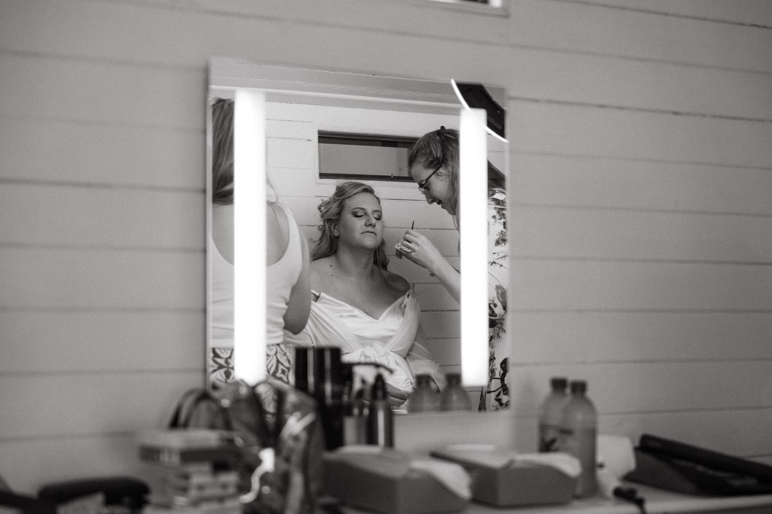 A black and white reflection in a vanity mirror shows someone getting ready amongst beauty products and cosmetics.