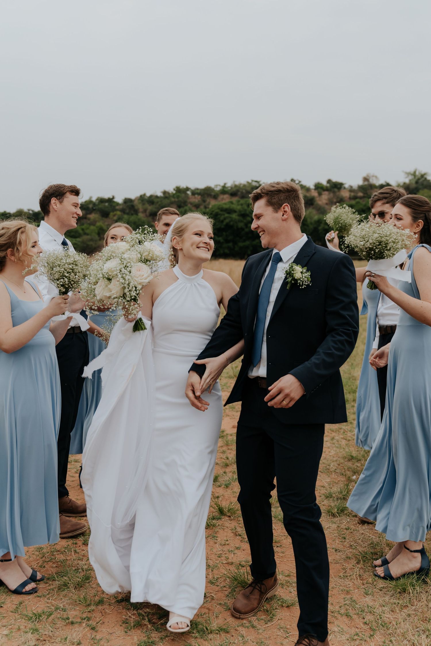 Wedding party in light blue bridesmaid dresses and dark suits dance joyfully outdoors on a sunny day.