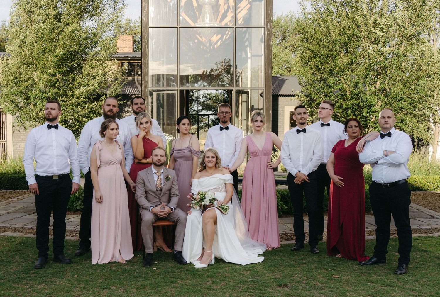 Wedding party poses together outdoors with trees and modern building facade in the background.
