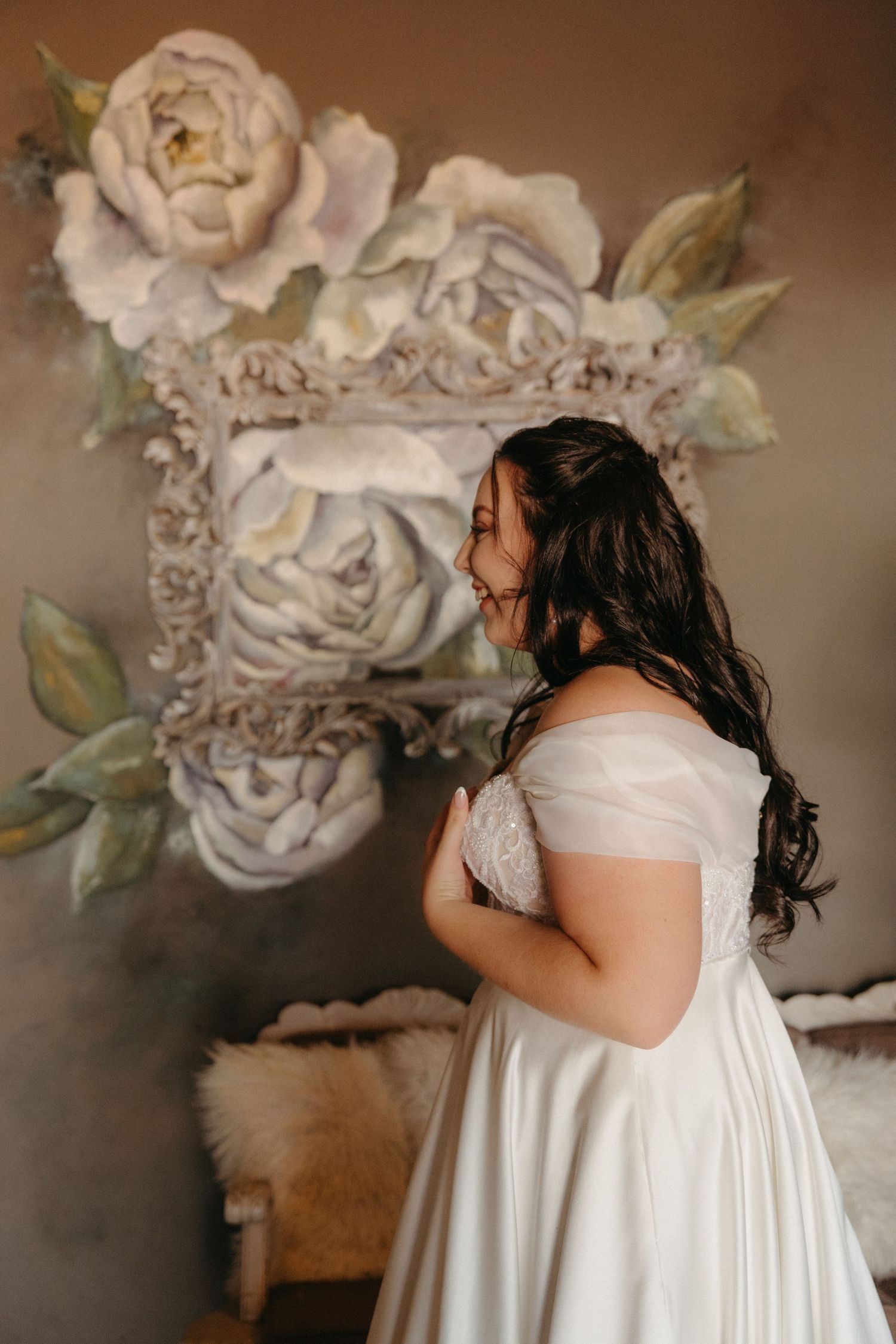 Side profile of a figure in a flowing white wedding gown posed in front of a wall with painted floral artwork.