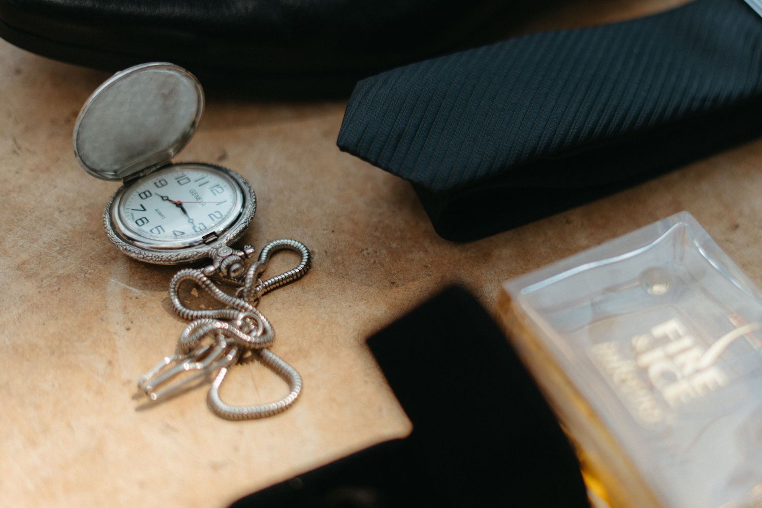 Vintage pocket watch with keys on a leather surface next to a black tie and plastic case.