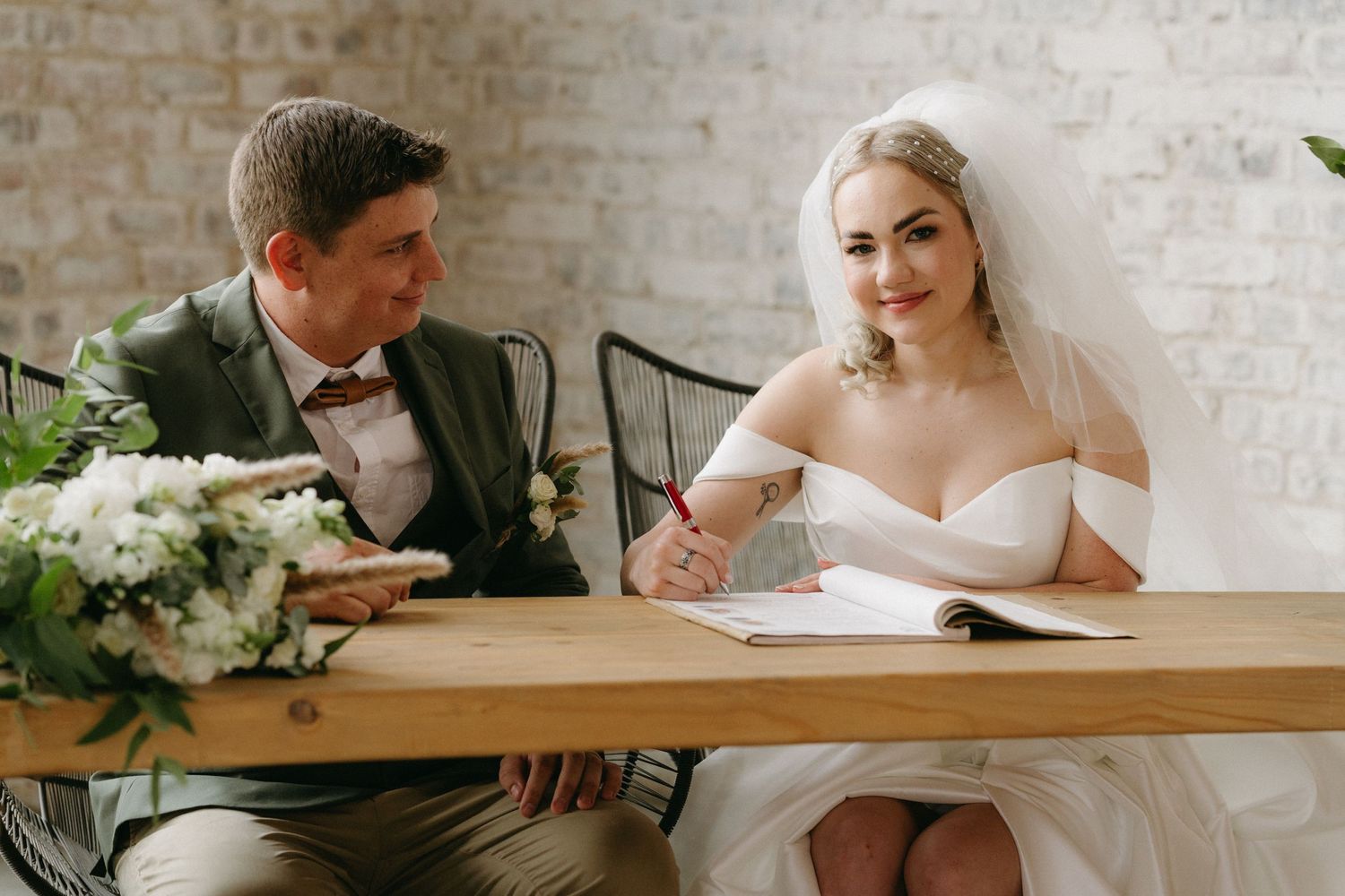 Couple signing marriage documents at wooden table decorated with white floral arrangements in bright indoor venue.