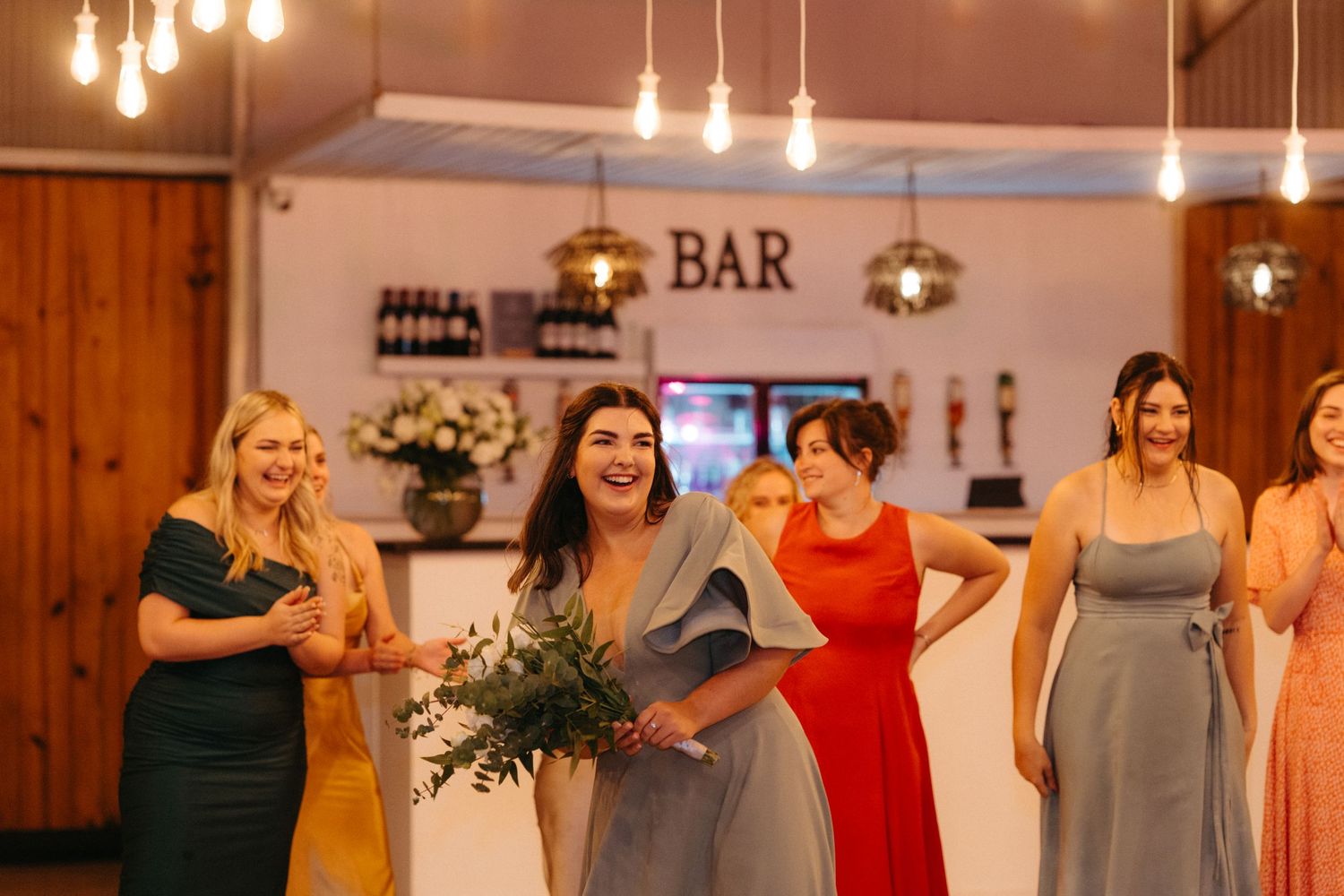 Bridesmaids in elegant dresses gather at a rustic bar venue with warm lighting and wooden accents.