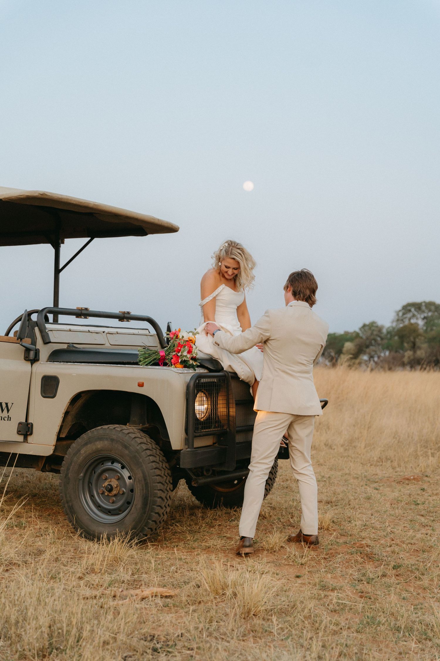Safari-style wedding photos with vintage Land Rover in golden field at dusk.