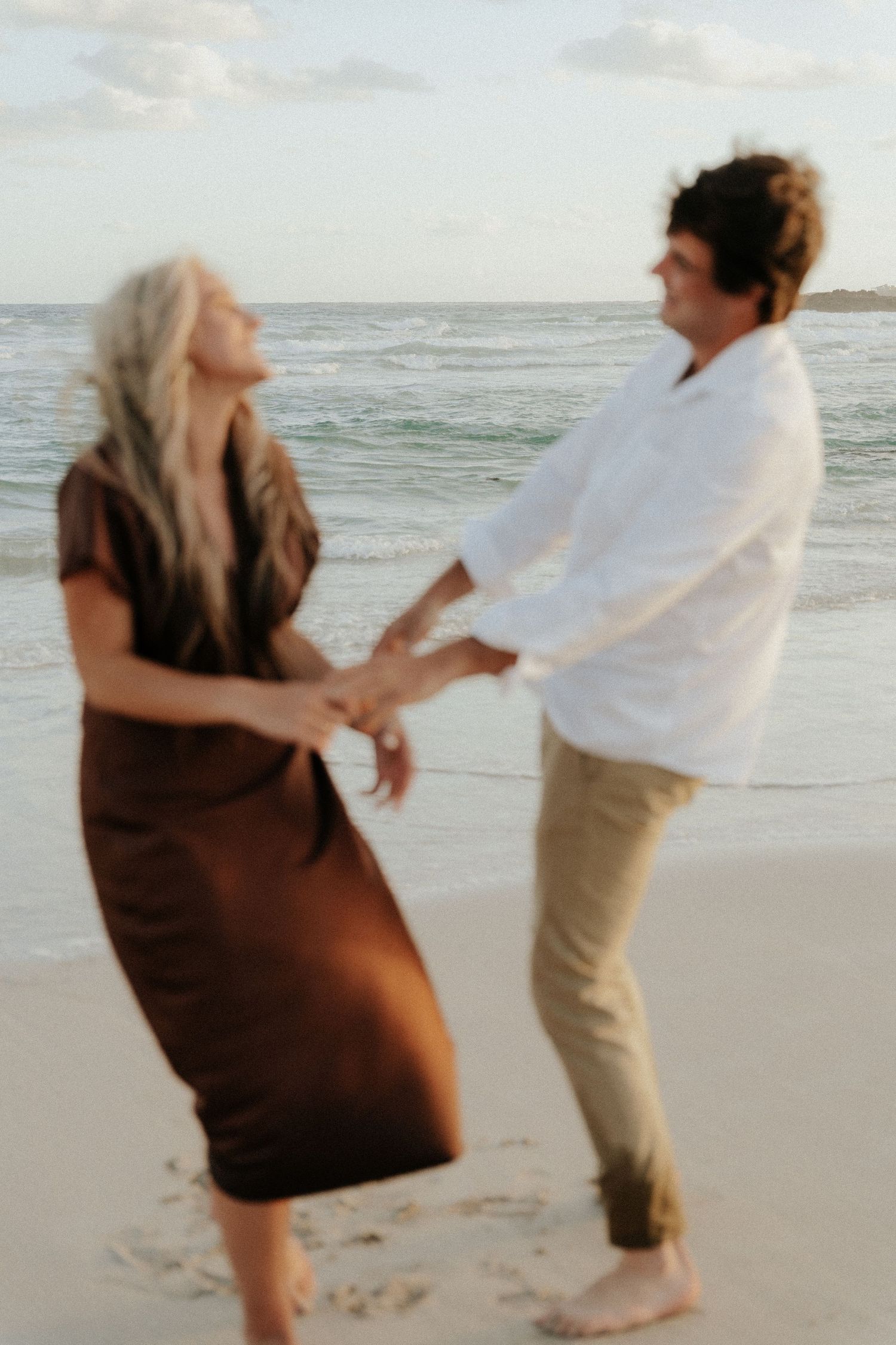 Couple dancing joyfully on a beach at sunset, woman in brown dress and man in white shirt holding hands.