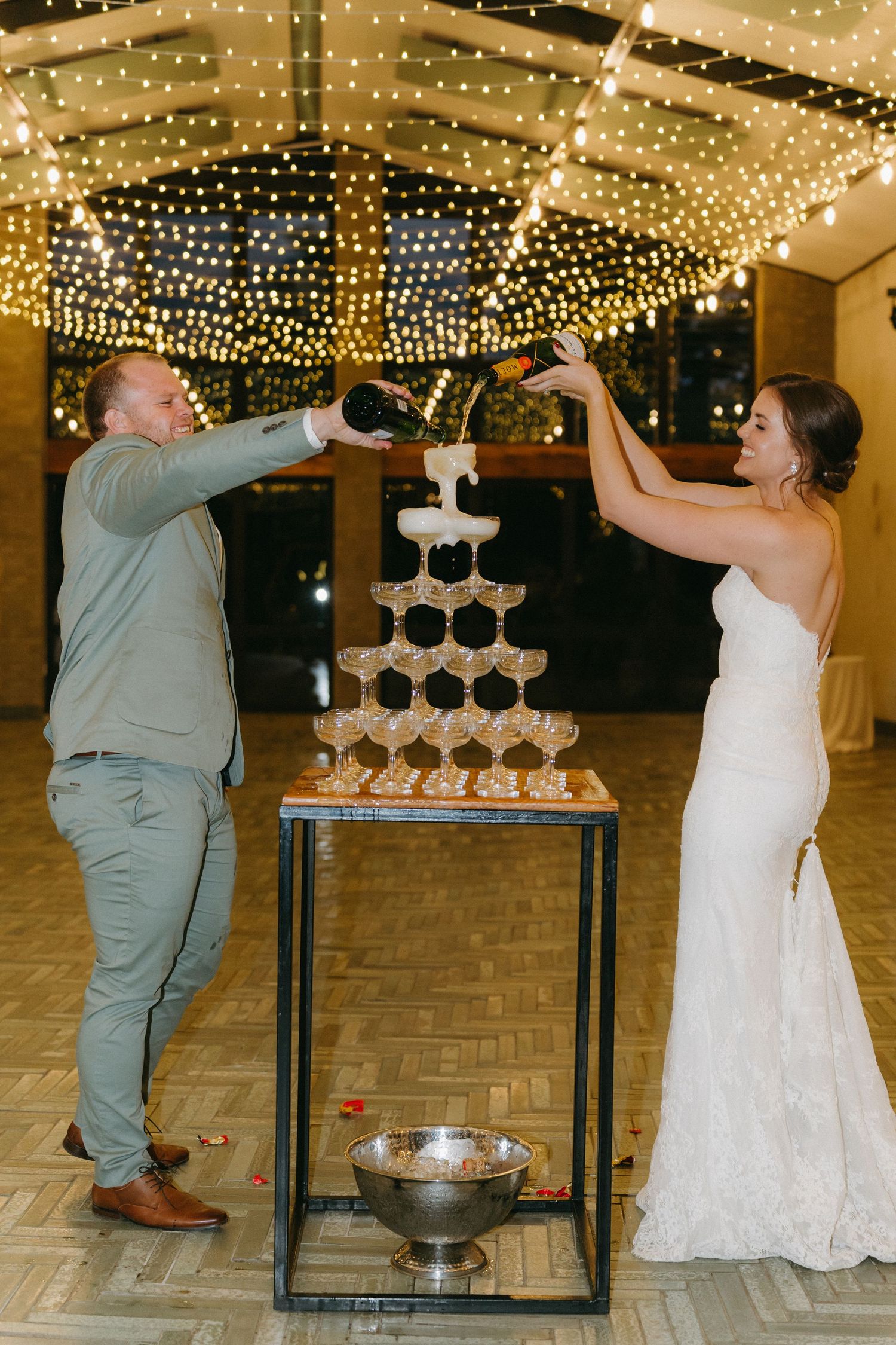 Wedding couple pouring champagne into tower of glasses under sparkling ceiling lights.