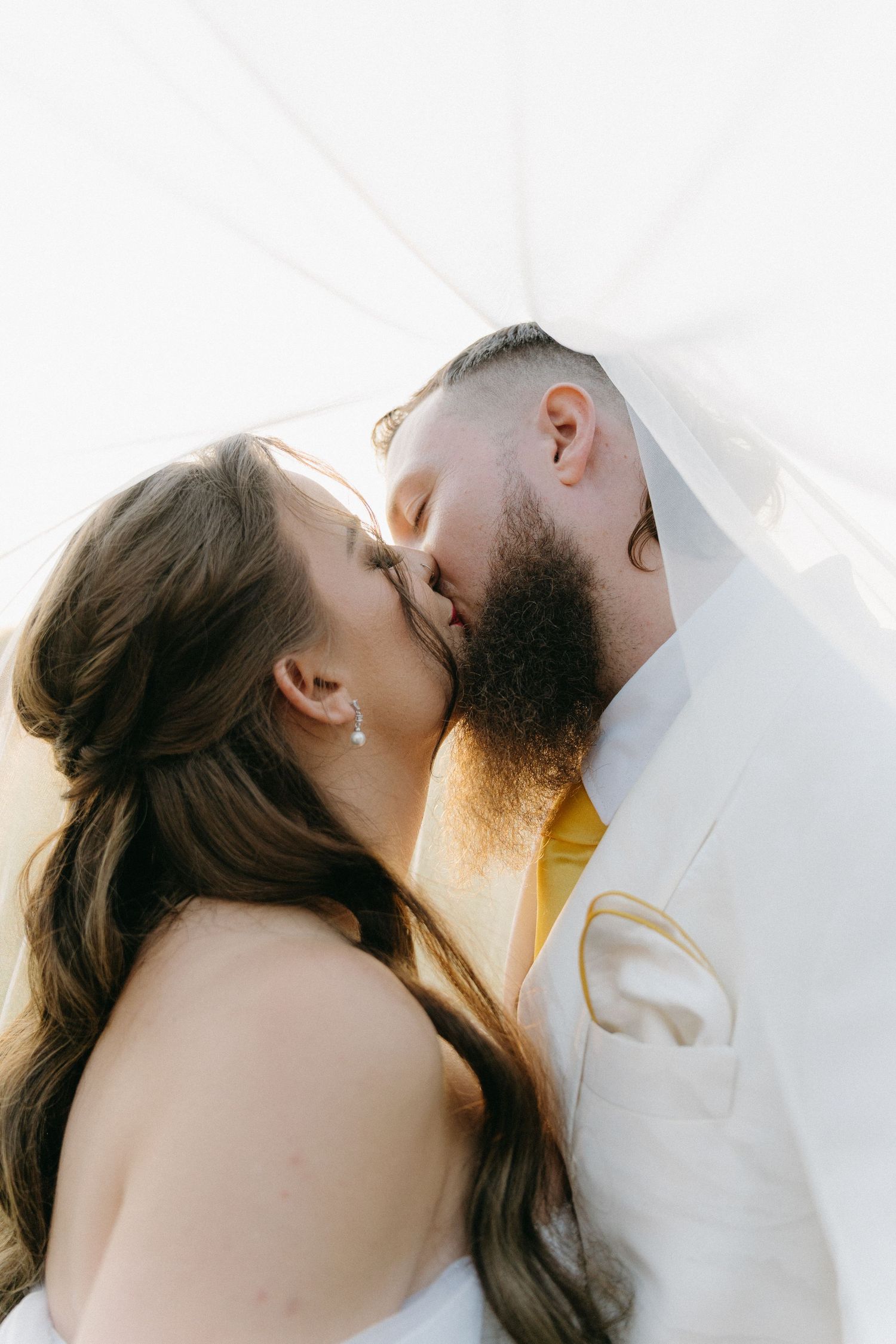 Close-up romantic moment between newlyweds in white attire against bright sunlight.