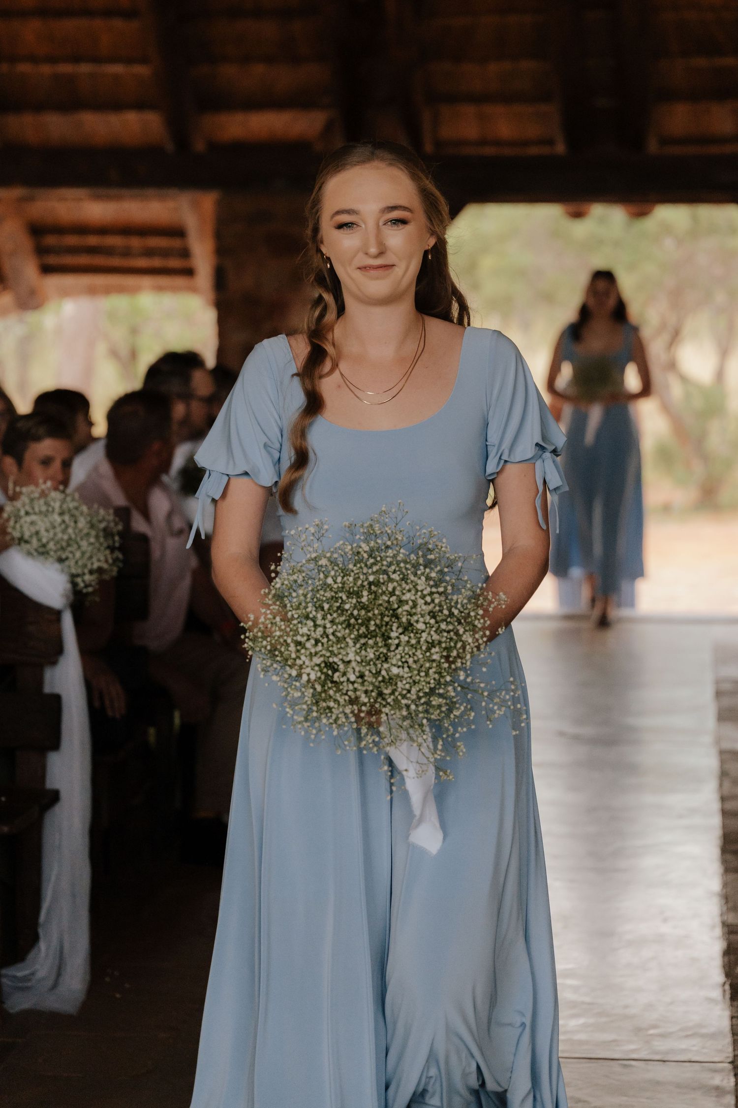 Bridesmaids in light blue dresses carry baby's breath bouquets while standing in a rustic venue.