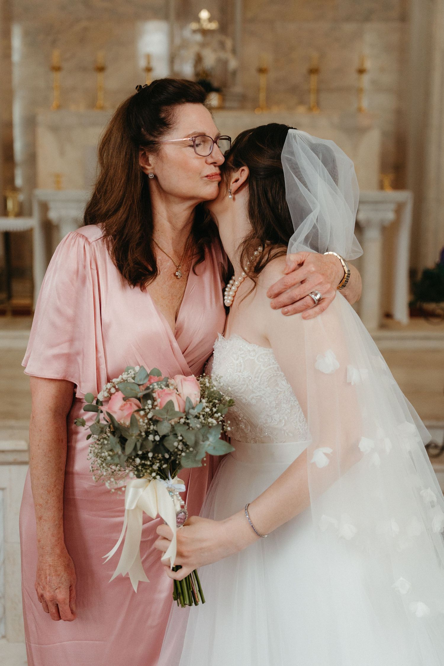 A tender moment captured at a wedding ceremony with a pink-dressed woman and bride holding a pastel bouquet in a church setting.