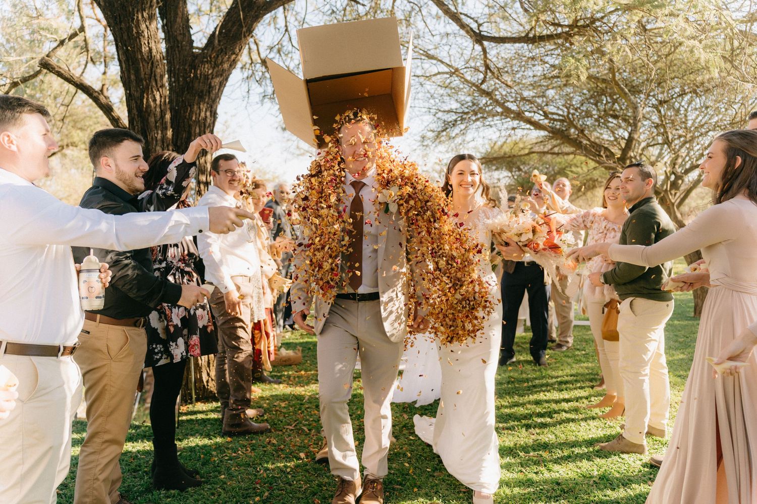 Wedding guests throw confetti from a box in celebration at an outdoor garden ceremony.