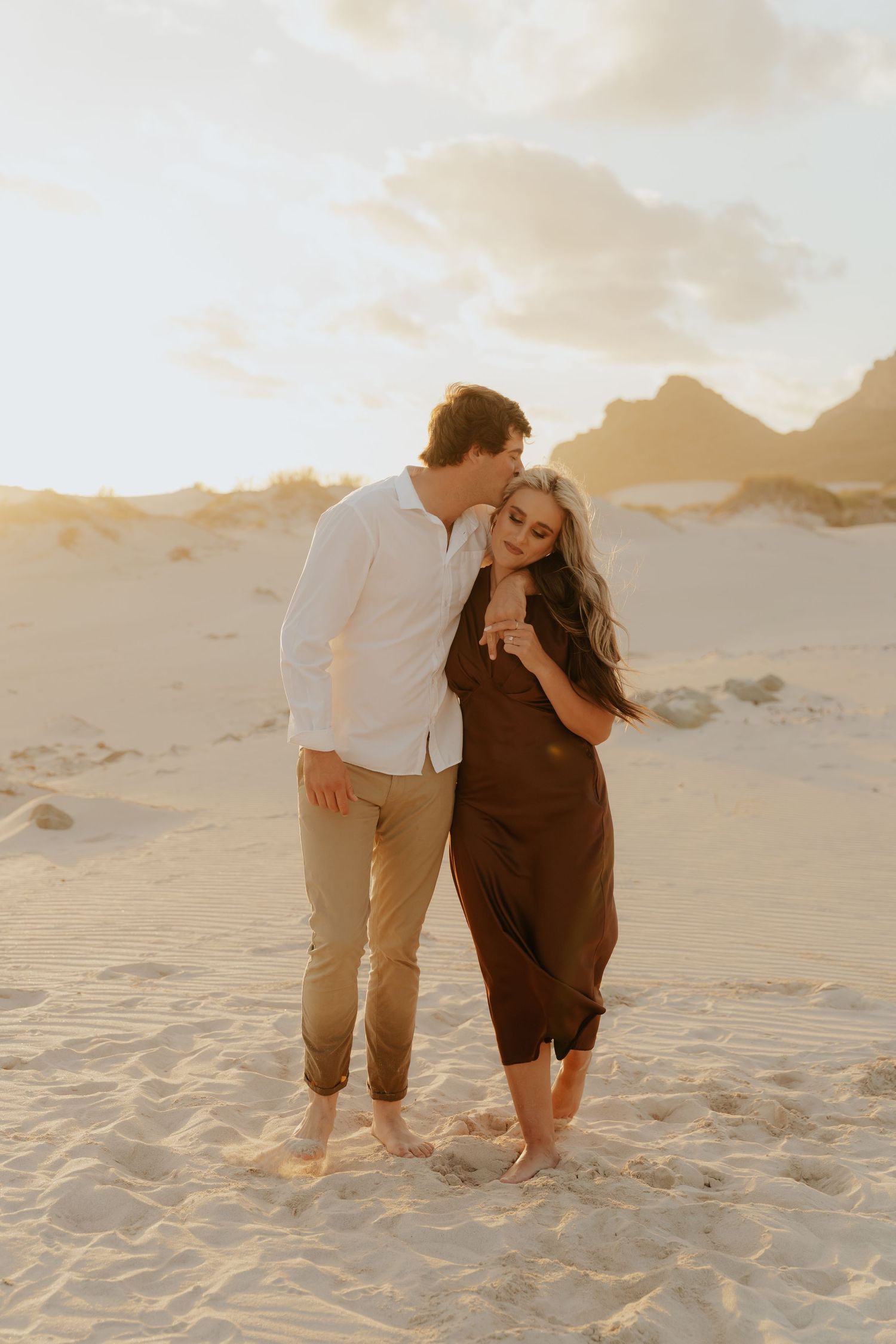 Couple embracing on sandy beach dunes at golden sunset, woman in brown dress, man in white shirt and khaki pants.