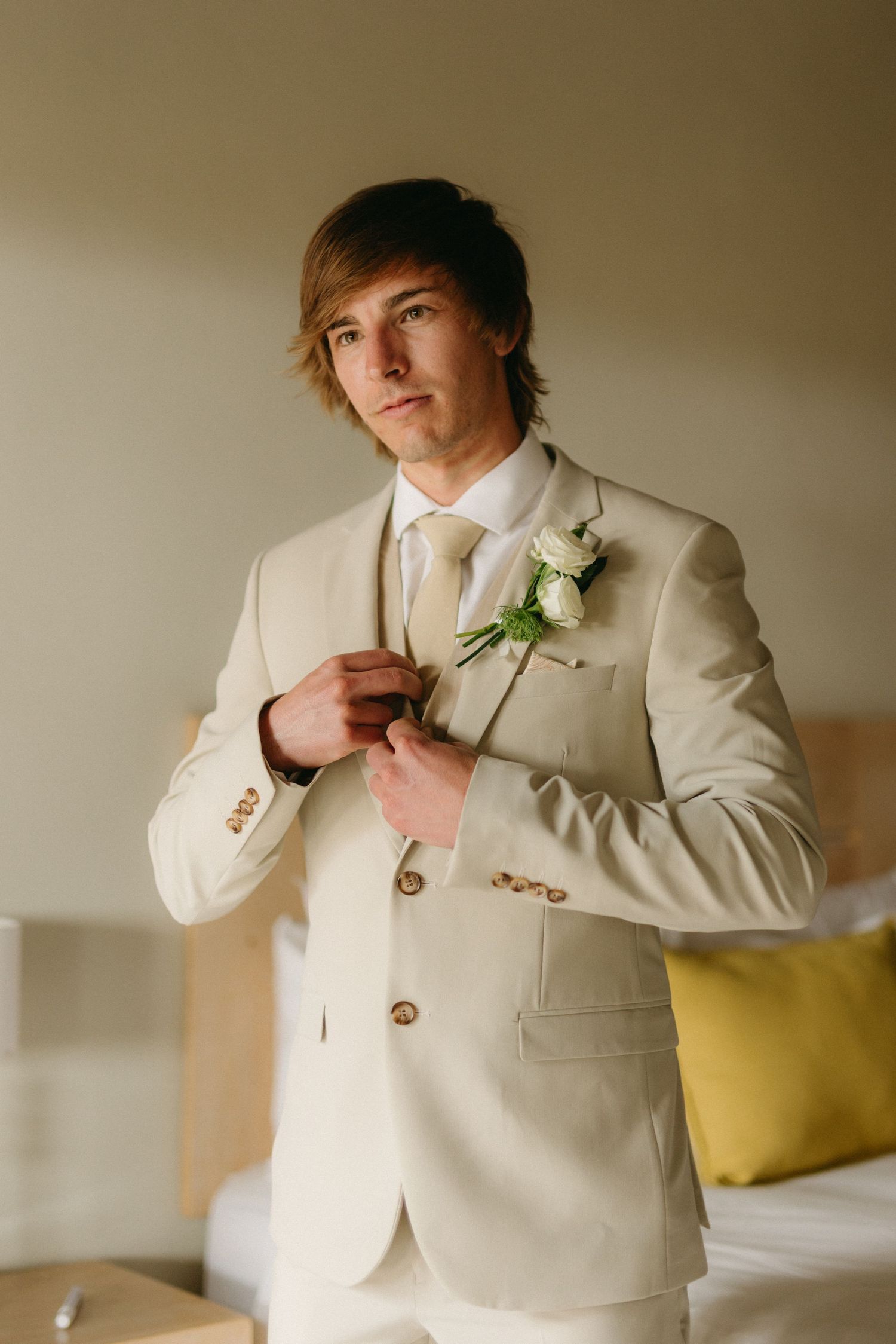 A formal cream-colored suit jacket with a white boutonniere being adjusted in preparation for a wedding.