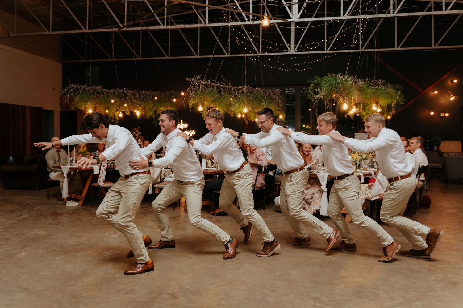 A line of groomsmen perform a synchronized dance routine at an indoor wedding reception venue with string lights overhead.