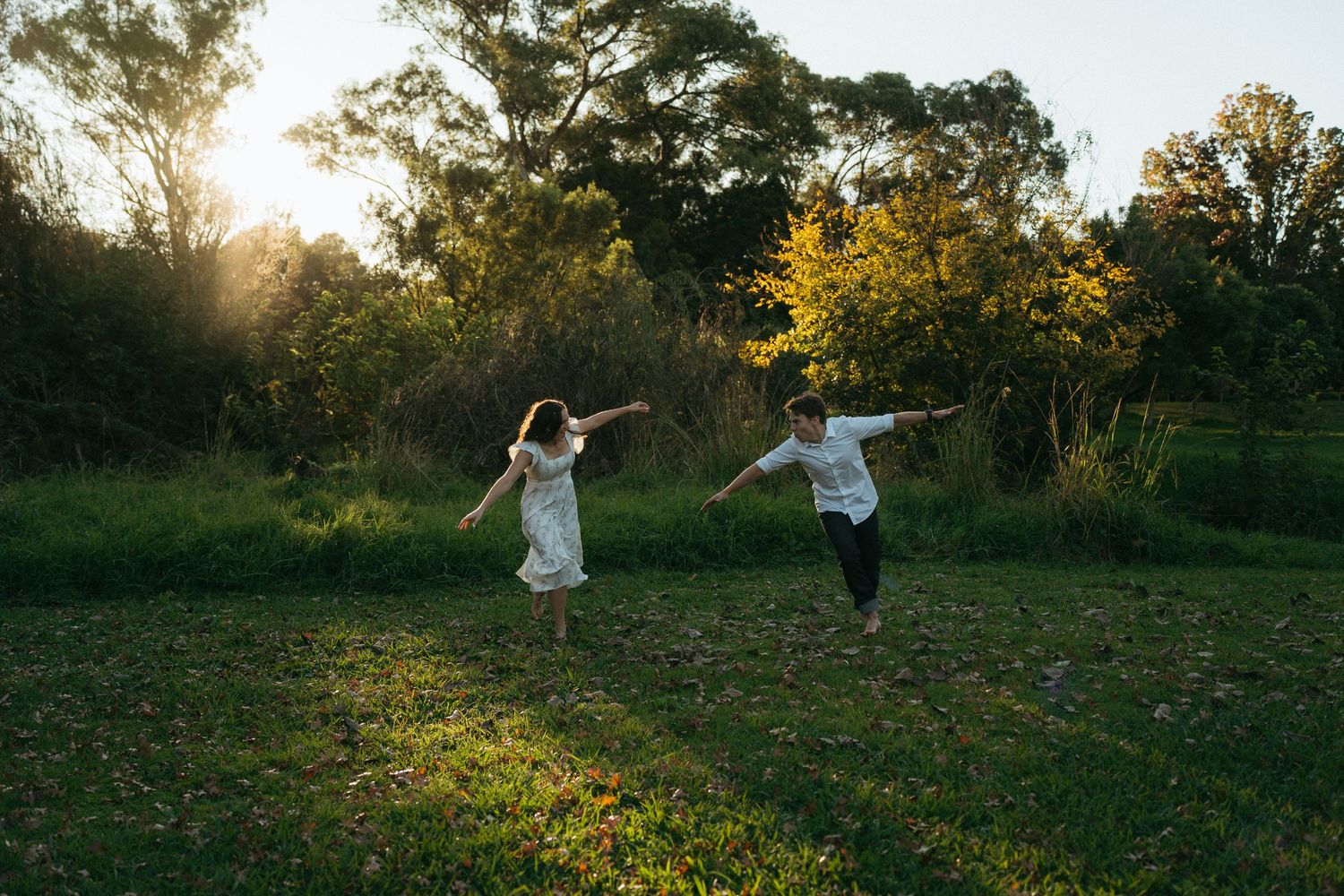 Two people in casual attire dance joyfully in a sunlit meadow during golden hour with trees in the background.