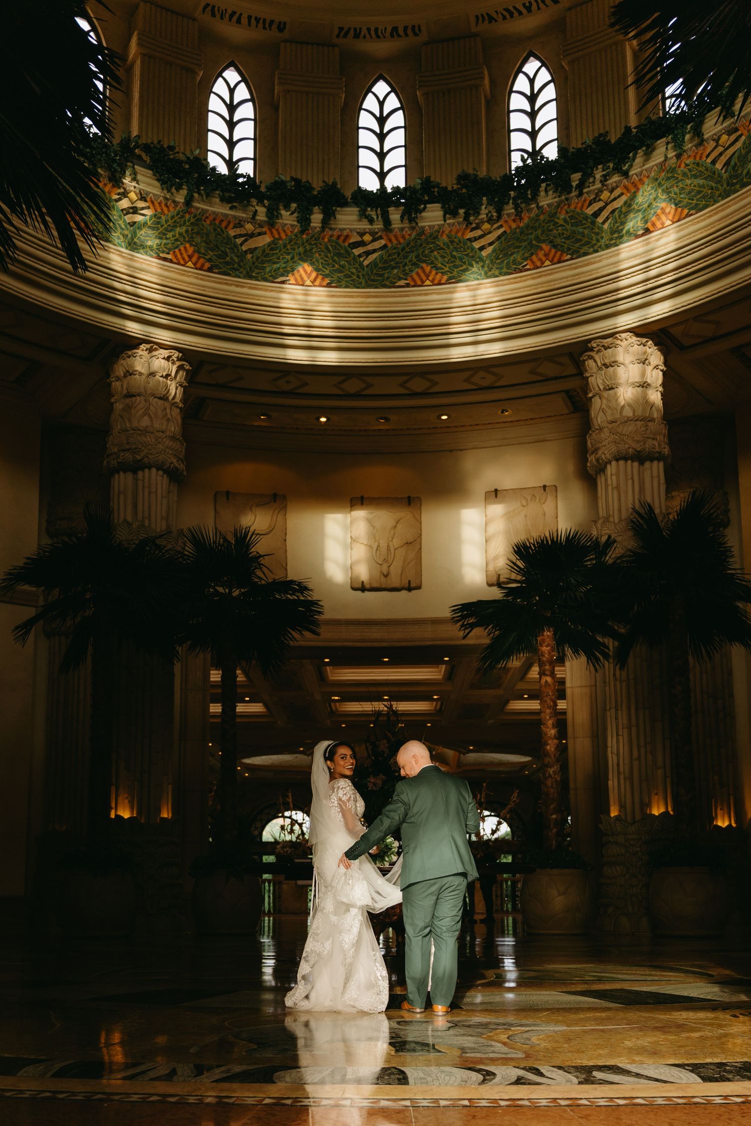 Newlyweds stand under grand domed ceiling with Gothic windows and palm decorations in dramatic lighting.